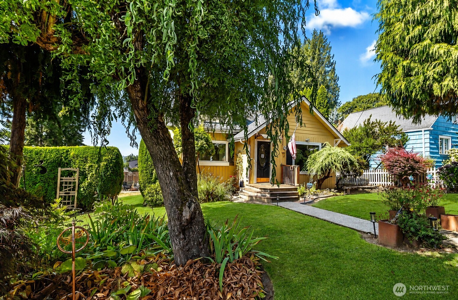 Yellow cottage with a front porch, framed by mature trees and a lush landscaped garden.