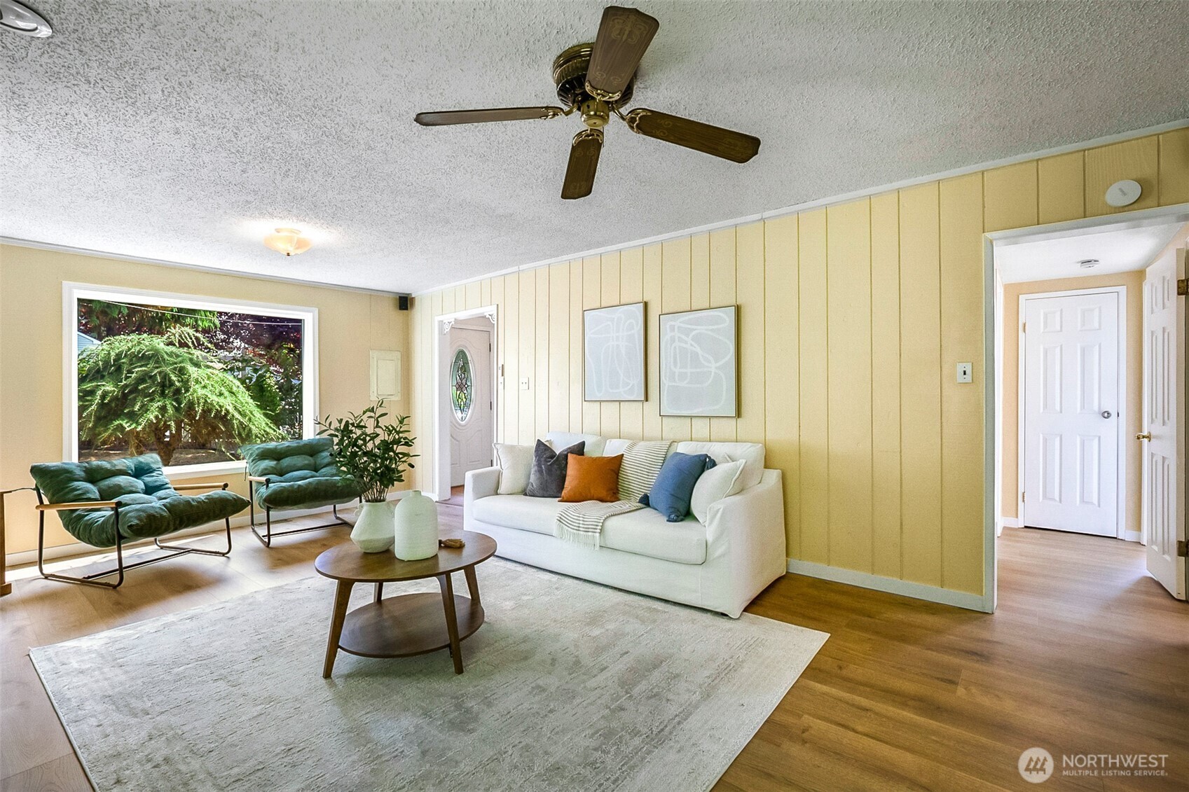 Living room with a white sofa, teal chairs, yellow paneled walls, and a large window overlooking greenery.