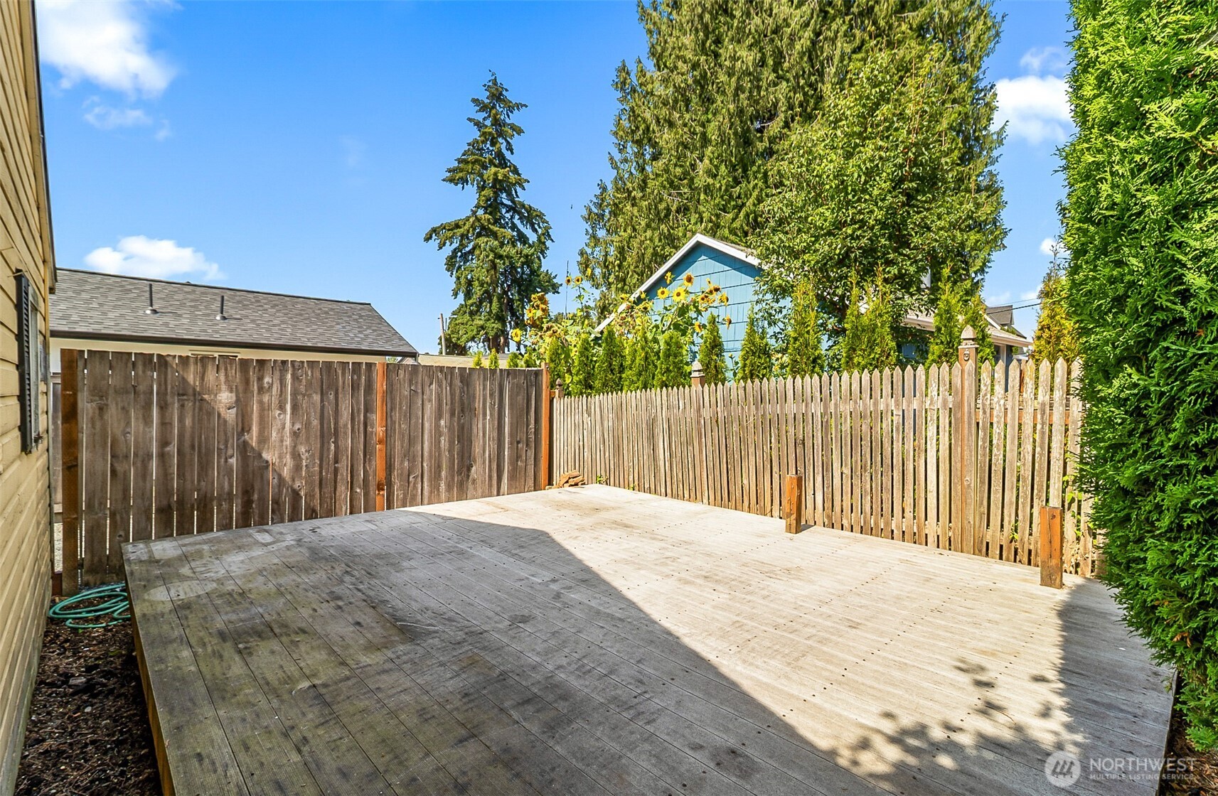 Spacious wooden backyard deck enclosed by a tall fence, with evergreens and sunflowers beyond.