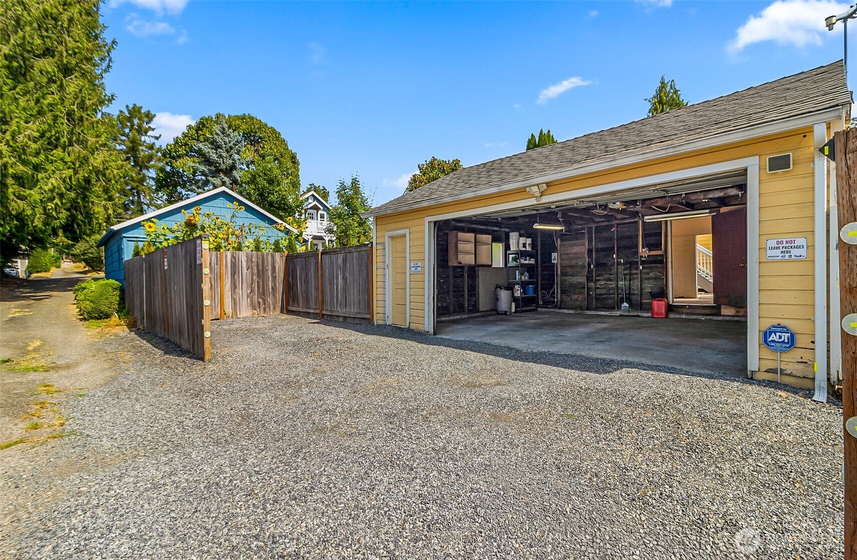 Yellow garage with open workshop door beside a broad gravel driveway and fenced blue outbuilding.