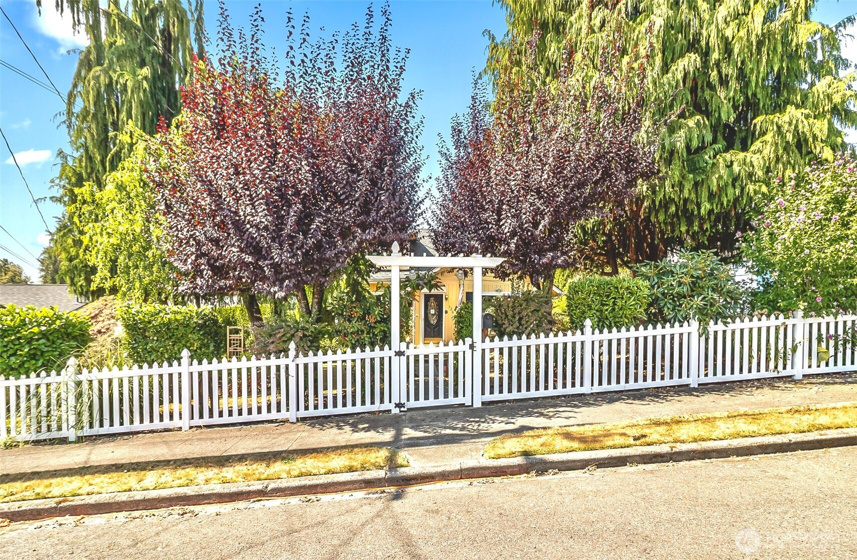 Home behind a white picket fence and arbor, framed by mature trees and garden landscaping.