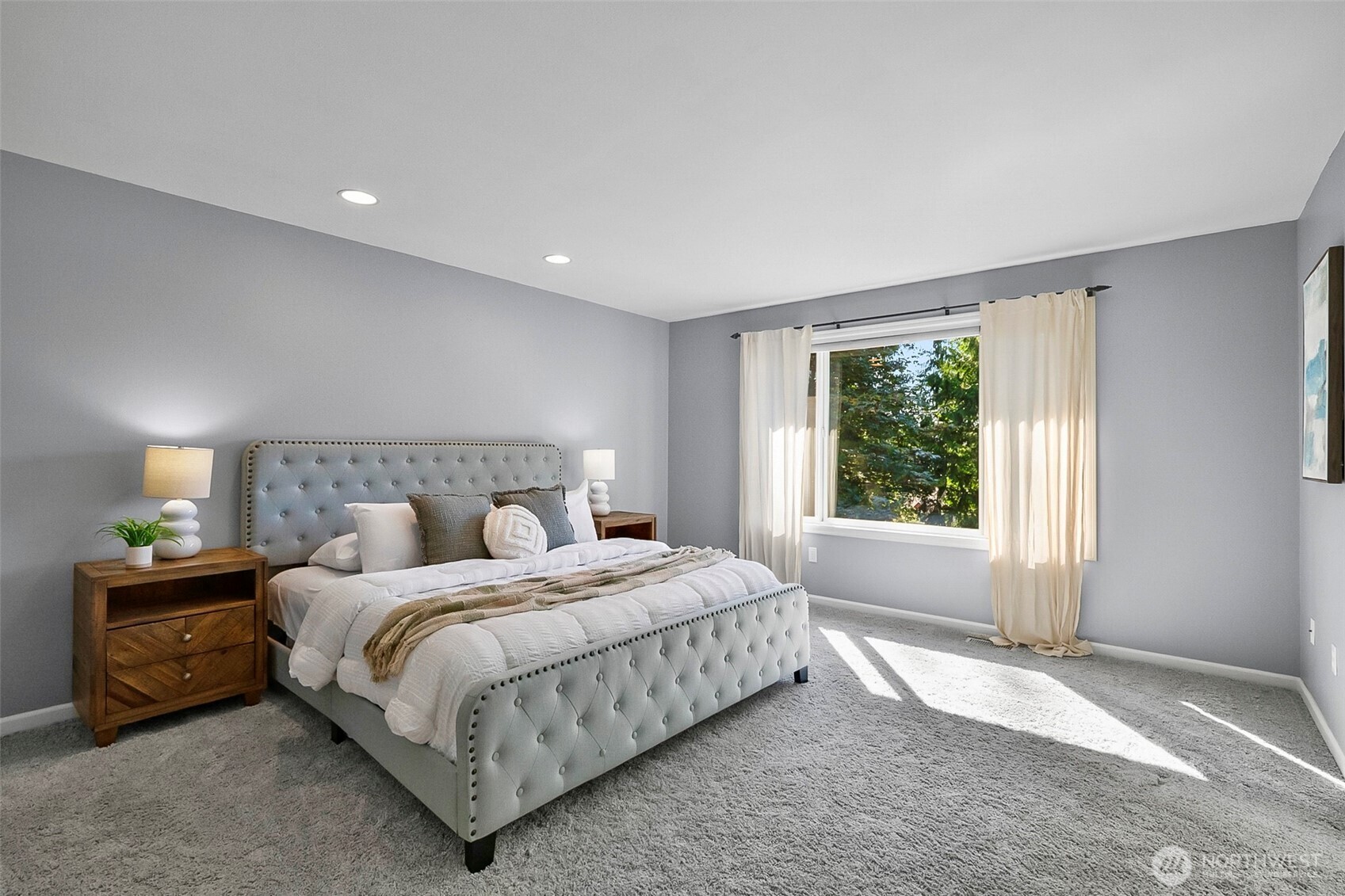 Staged bedroom with a gray tufted bed, white bedding, and sunlight through a curtained window.