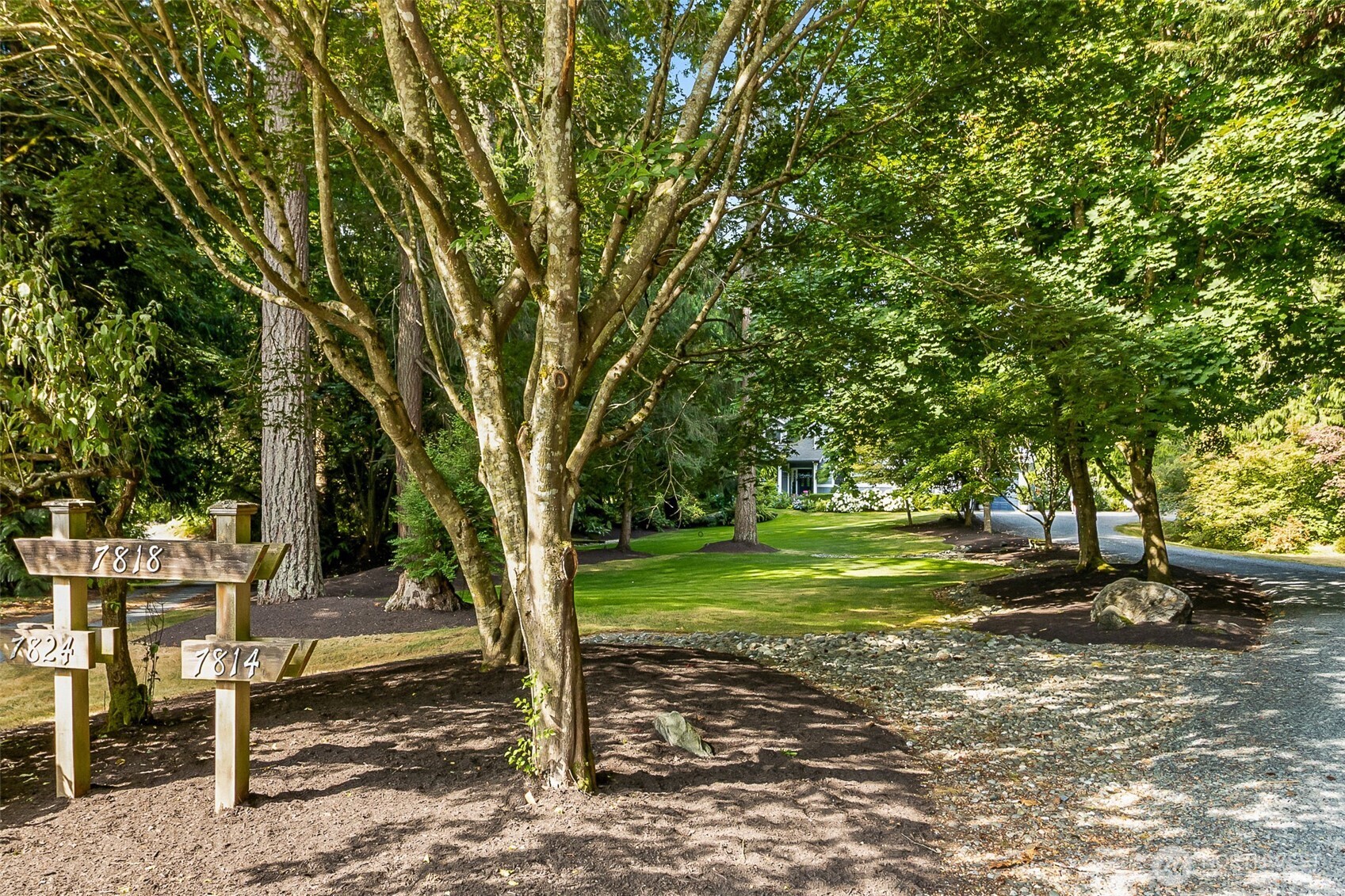 Tree-lined property entrance with a landscaped lawn, gravel driveway, and wooden 7818 address marker.