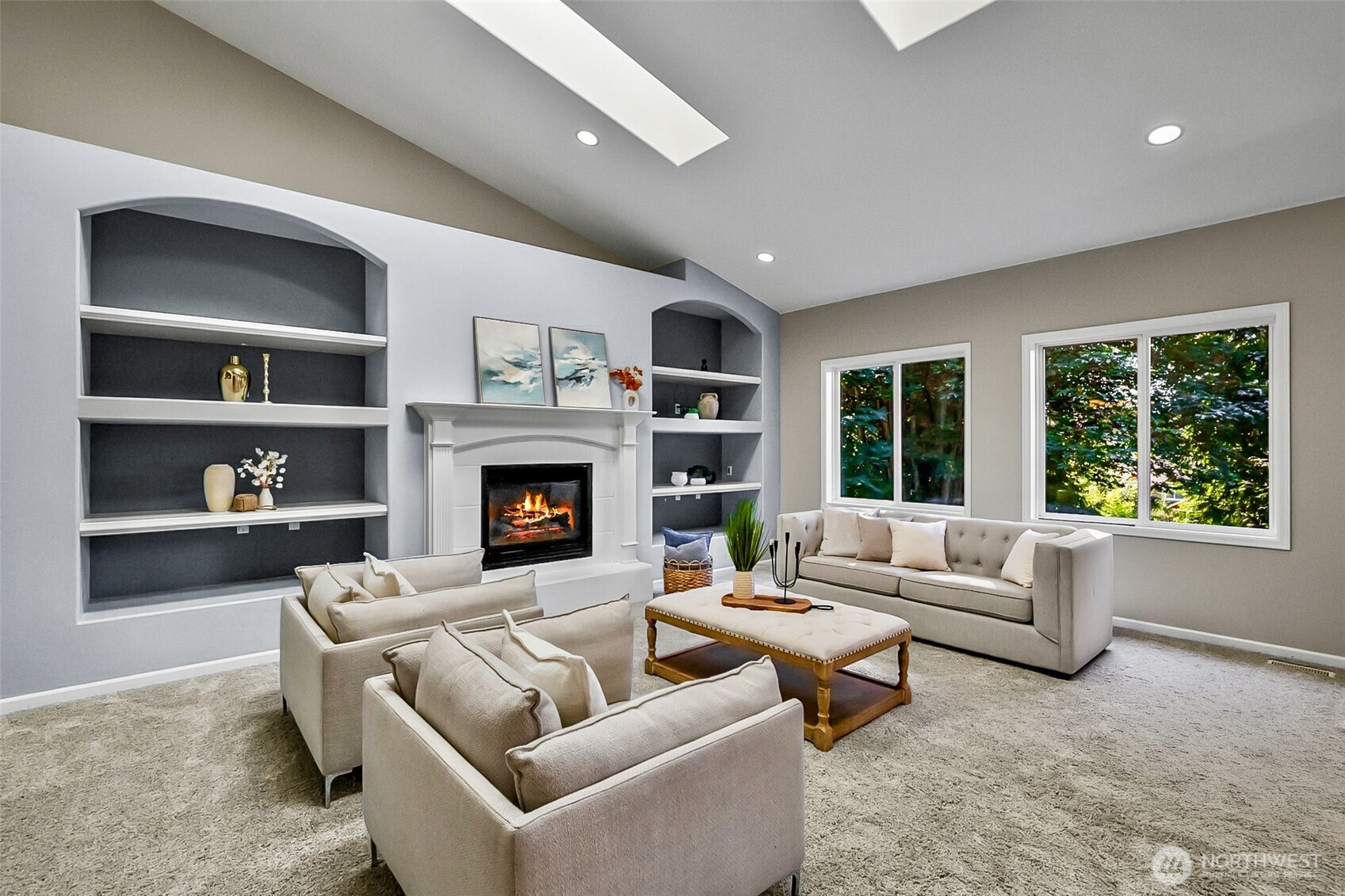 Staged living room with a vaulted ceiling, built-in shelves, a lit fireplace, and large windows overlooking trees.