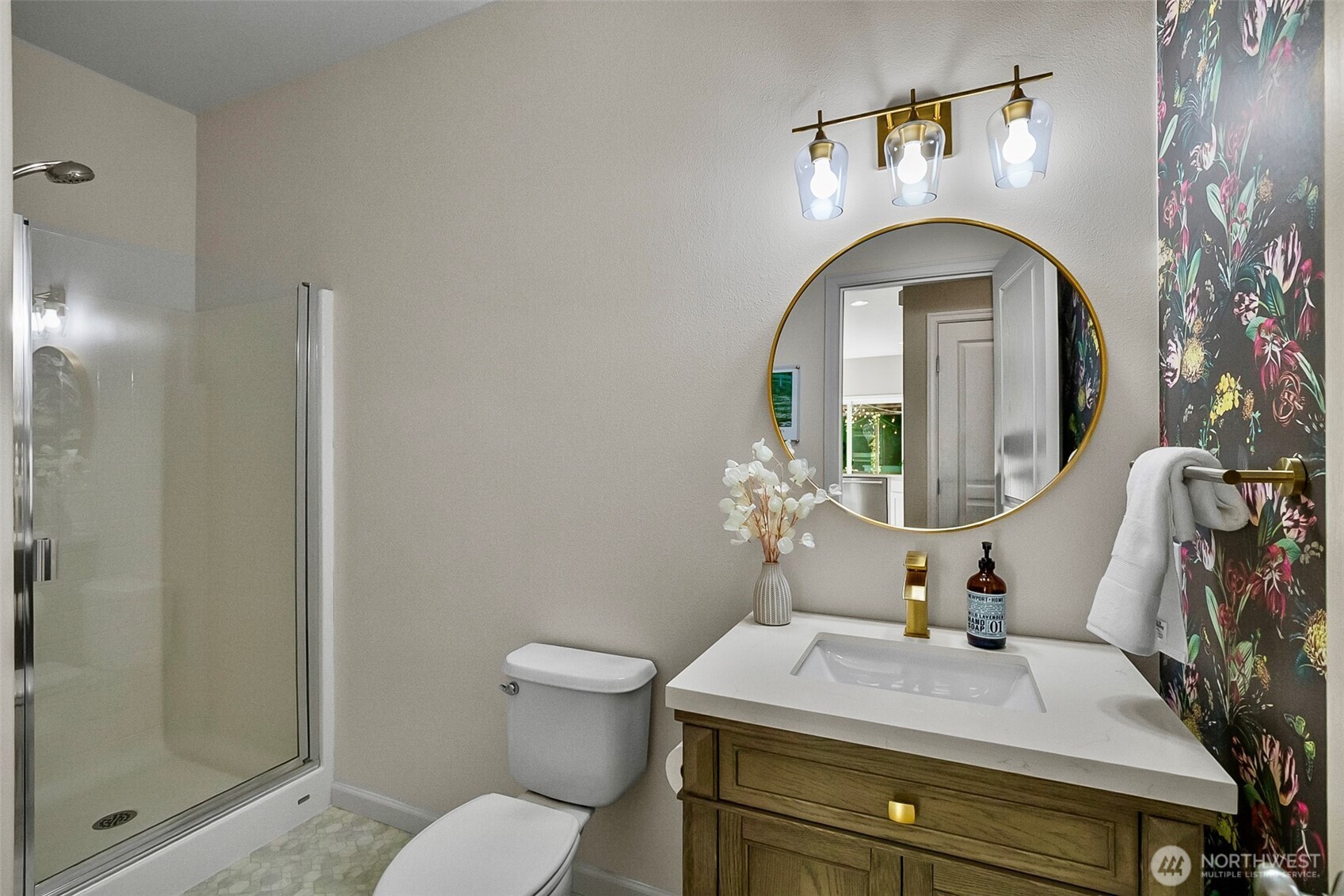 Bathroom with glass shower, wood vanity, round gold-framed mirror, and dark floral wallpaper.