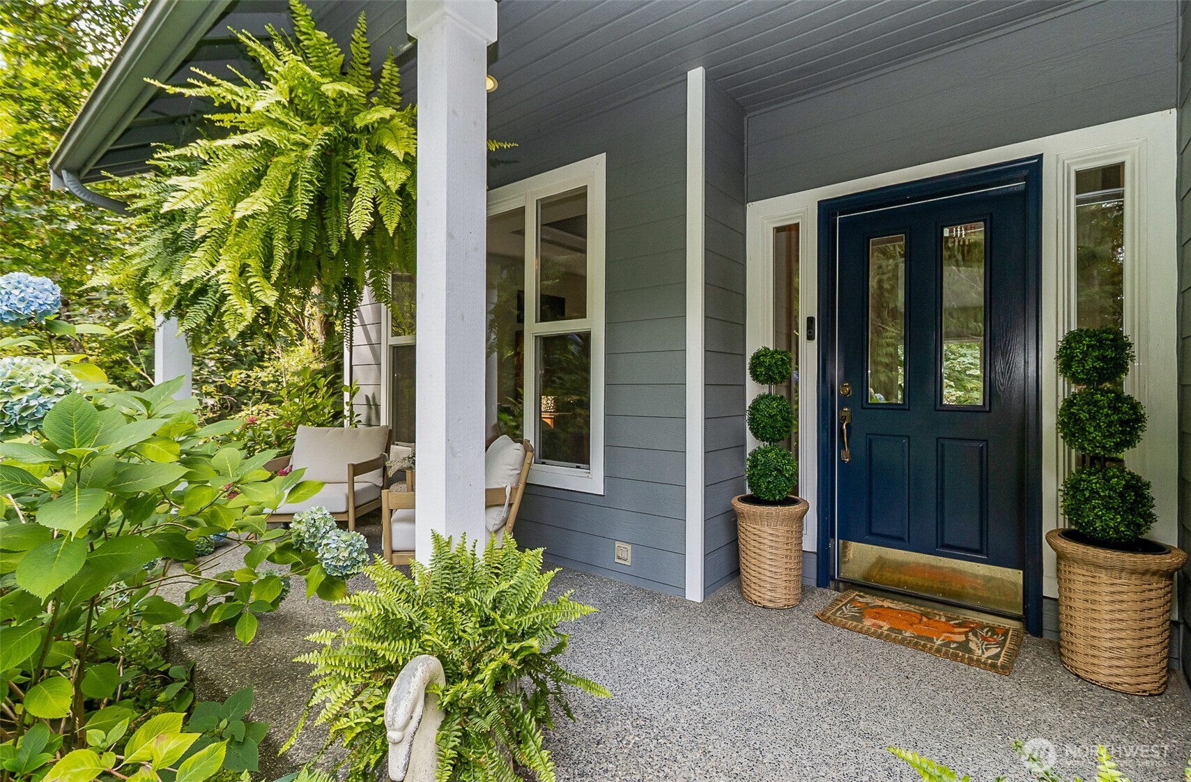 Gray home’s covered front porch with a dark blue door, cushioned chairs, and lush garden planting.