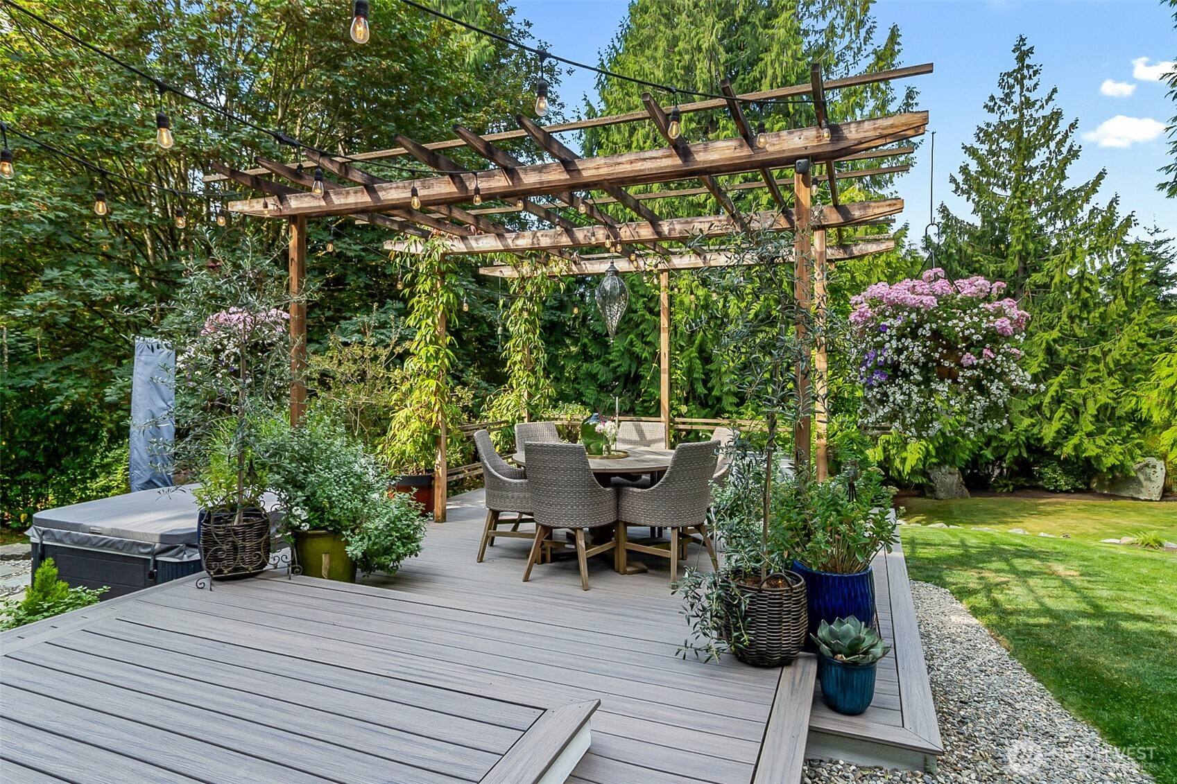 Outdoor deck with a pergola, string lights, dining set, and covered hot tub amid lush gardens.