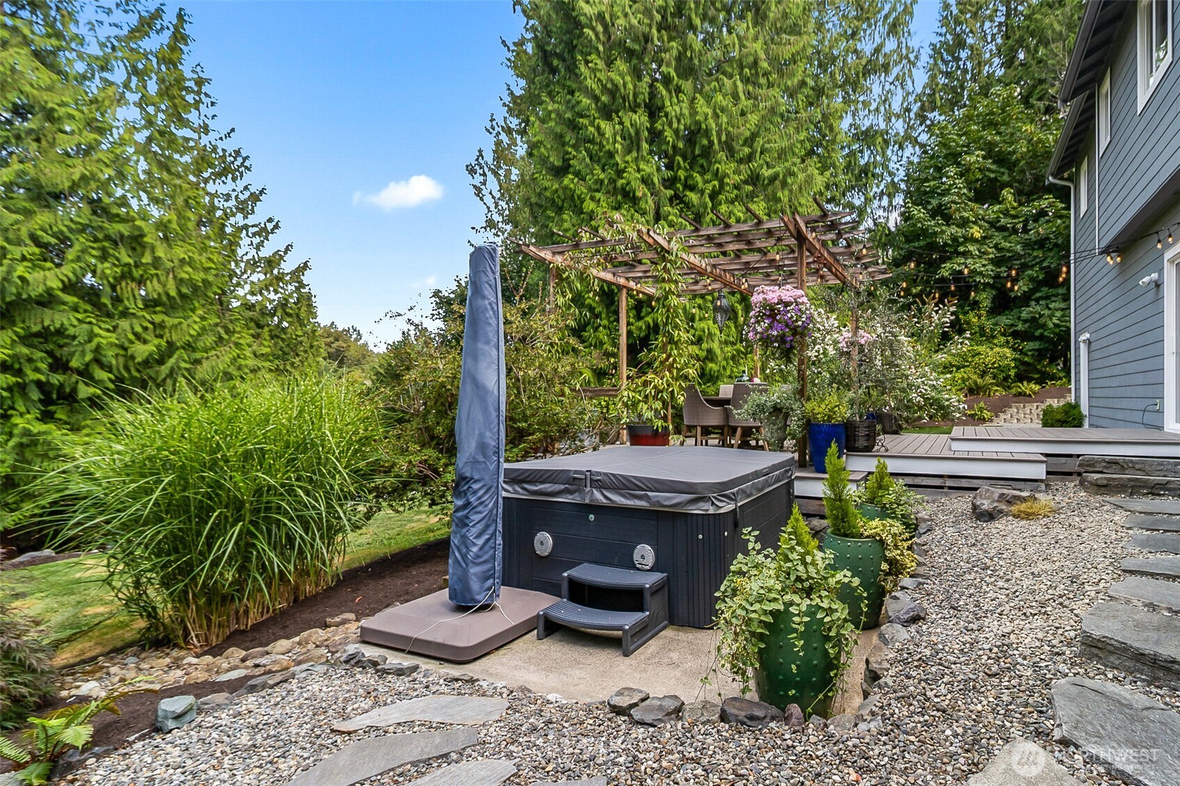 Backyard hot tub beside a pergola-covered dining area, deck, and flowering garden.