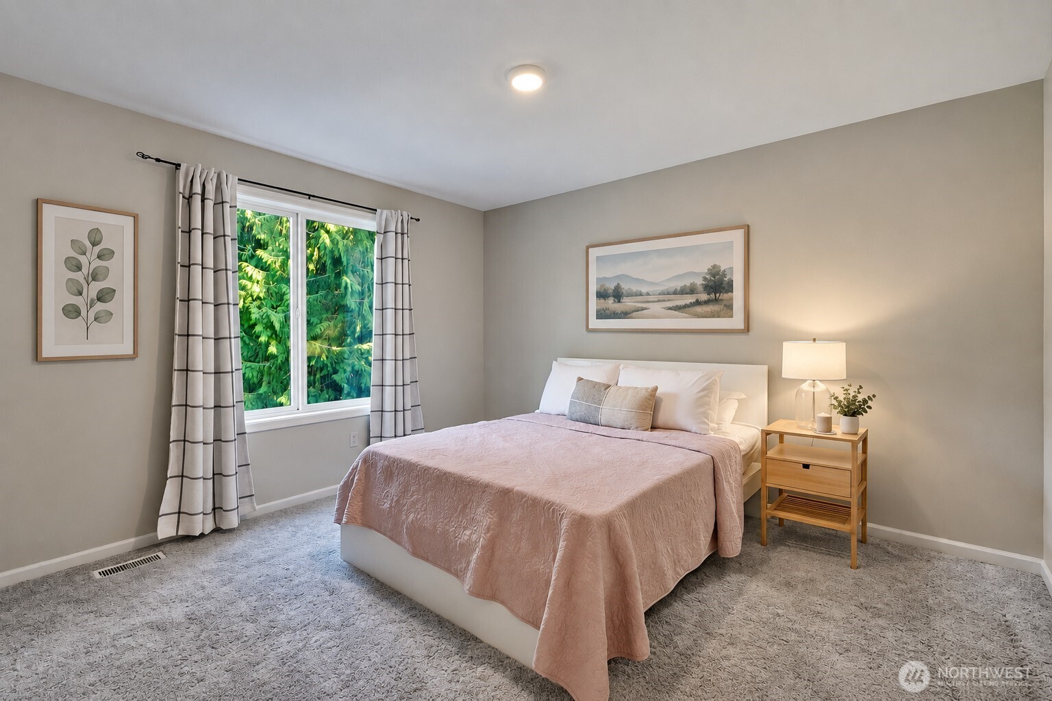 Bedroom with a pale pink quilted bed beside a window overlooking evergreen trees.