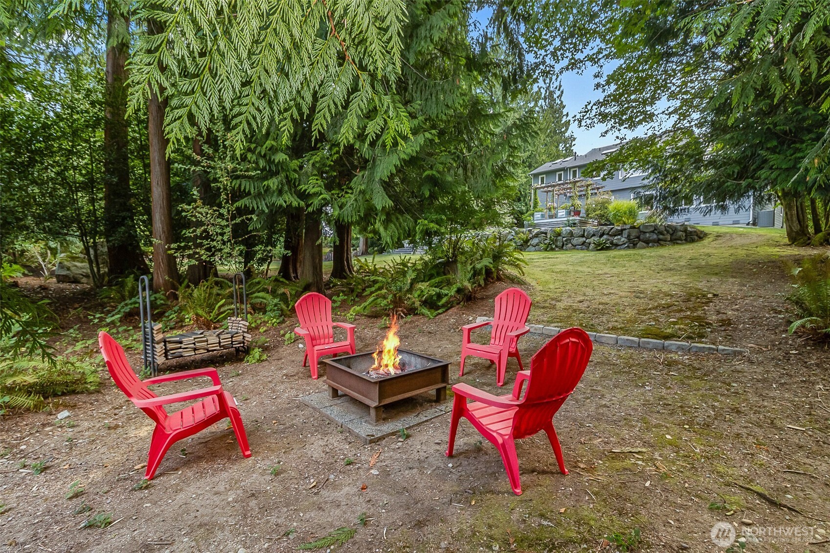 Wooded backyard fire pit surrounded by red Adirondack chairs beneath tall evergreen trees.