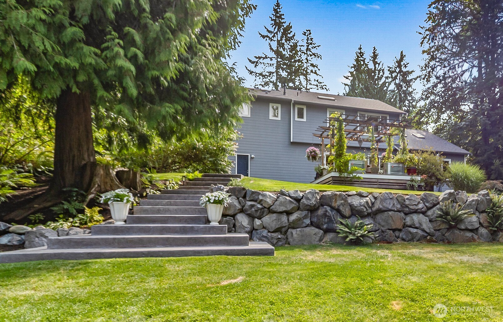 Gray two-story home with a pergola-covered deck, stone retaining wall, and broad garden steps.