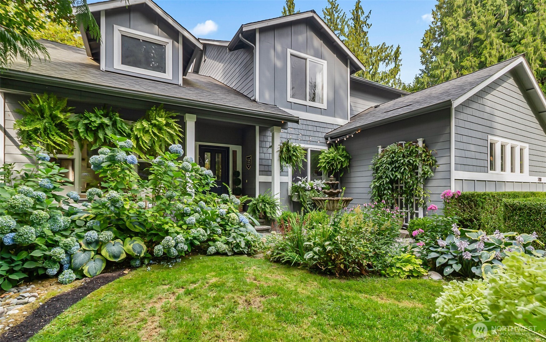 Gray two-story home with a covered entry, hanging ferns, and a lush hydrangea garden.