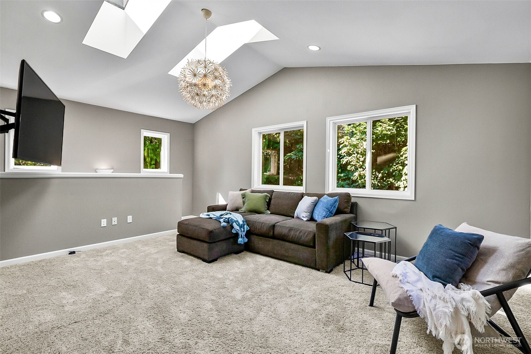 Furnished living room with vaulted ceiling, skylights, dark sectional, and windows overlooking greenery.