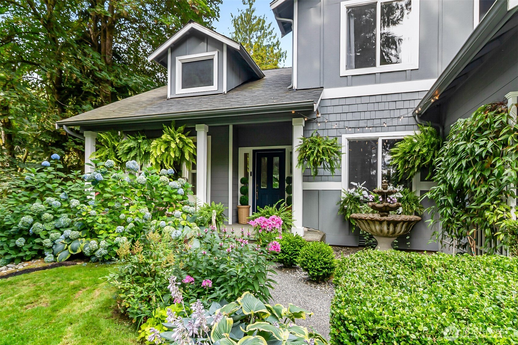 Gray two-story home with a covered entry, flowering hydrangeas, hanging ferns, and a tiered garden fountain.