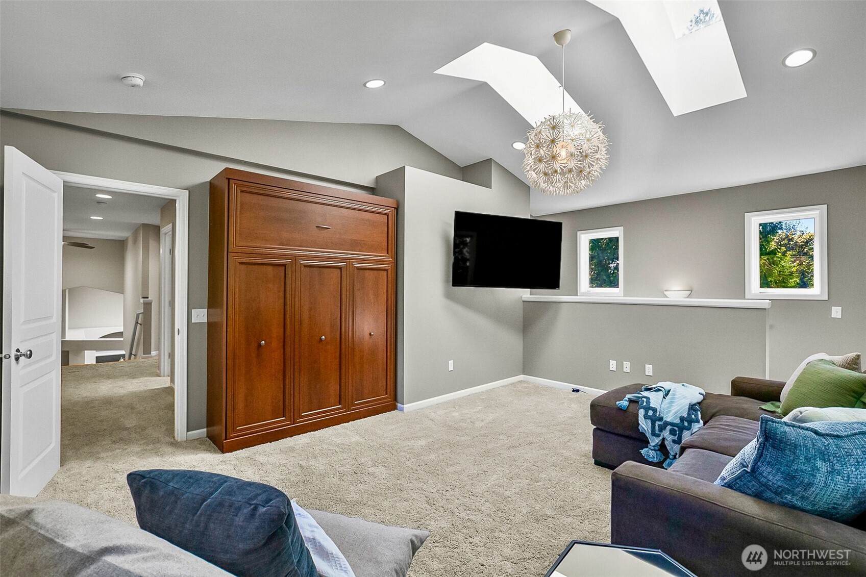 Furnished bonus room with a wall-mounted TV, wooden Murphy bed cabinet, skylights, and gray carpet.