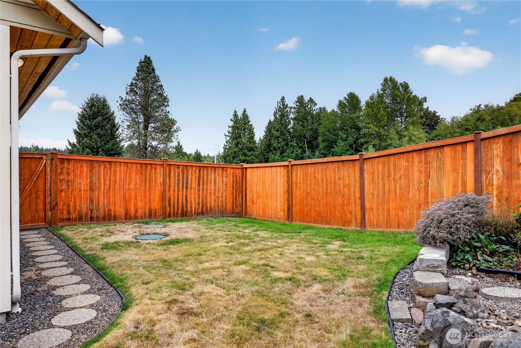 Fenced backyard with a patchy lawn, stepping-stone path, and rock garden beds.