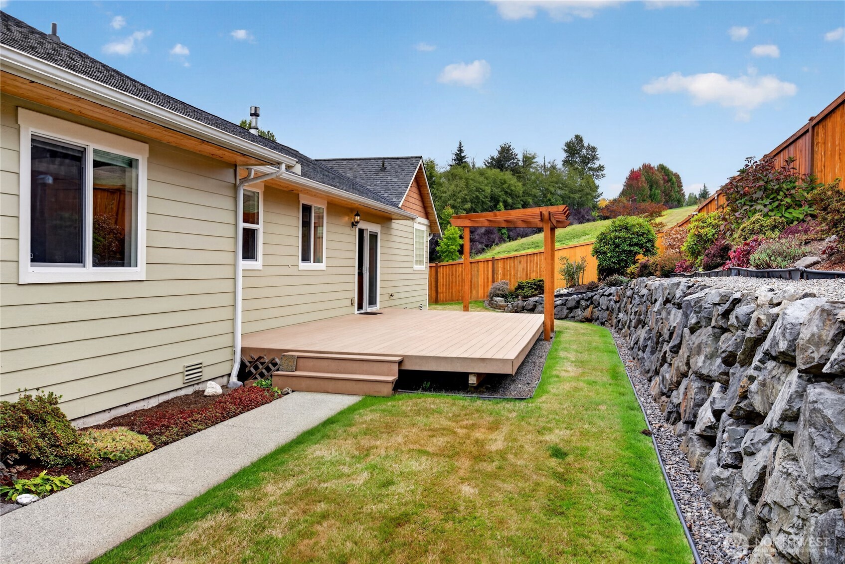 Backyard with a raised deck and pergola beside a landscaped lawn and stone retaining wall.