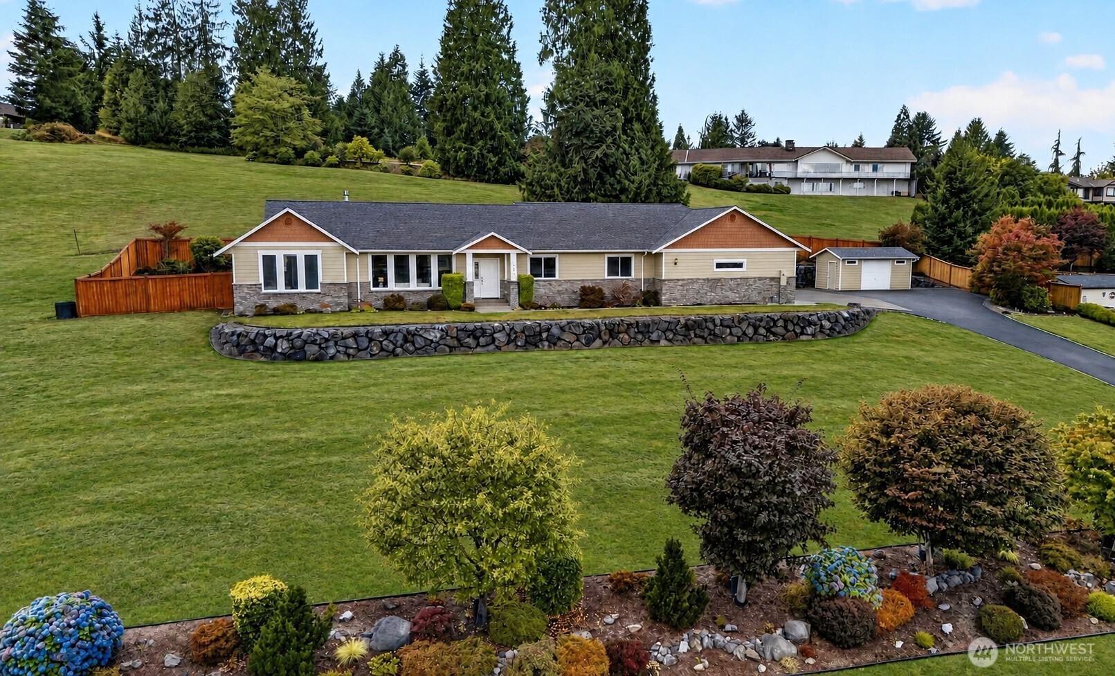 Single-story home with attached garage, broad sloping lawn, stone retaining wall, and evergreen trees.