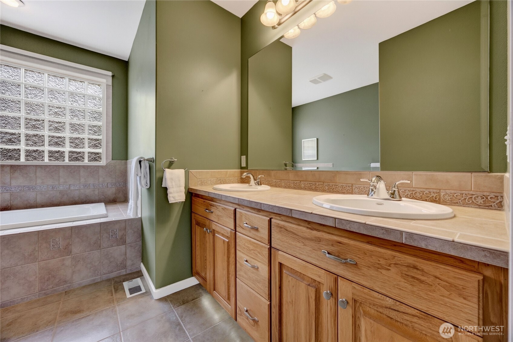 Bathroom with a double-sink wood vanity and tiled tub beneath a glass-block window.