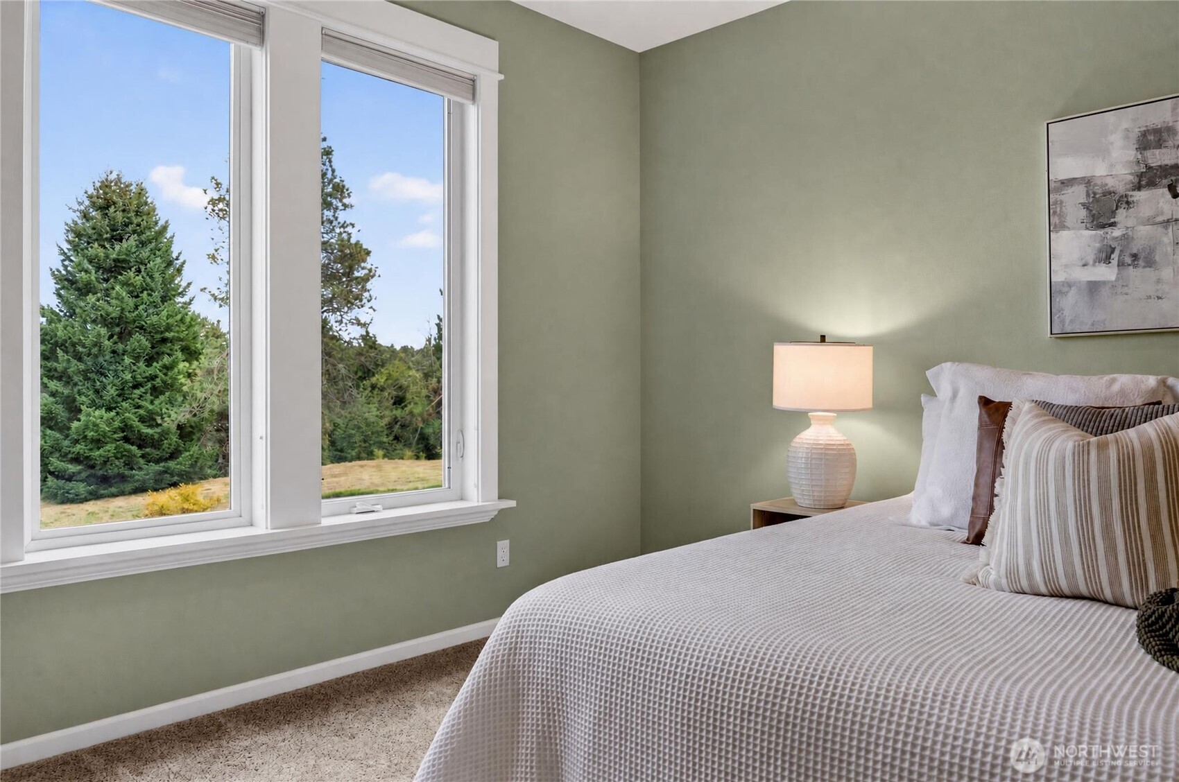 Staged bedroom with a white quilted bed and large windows overlooking evergreen trees.