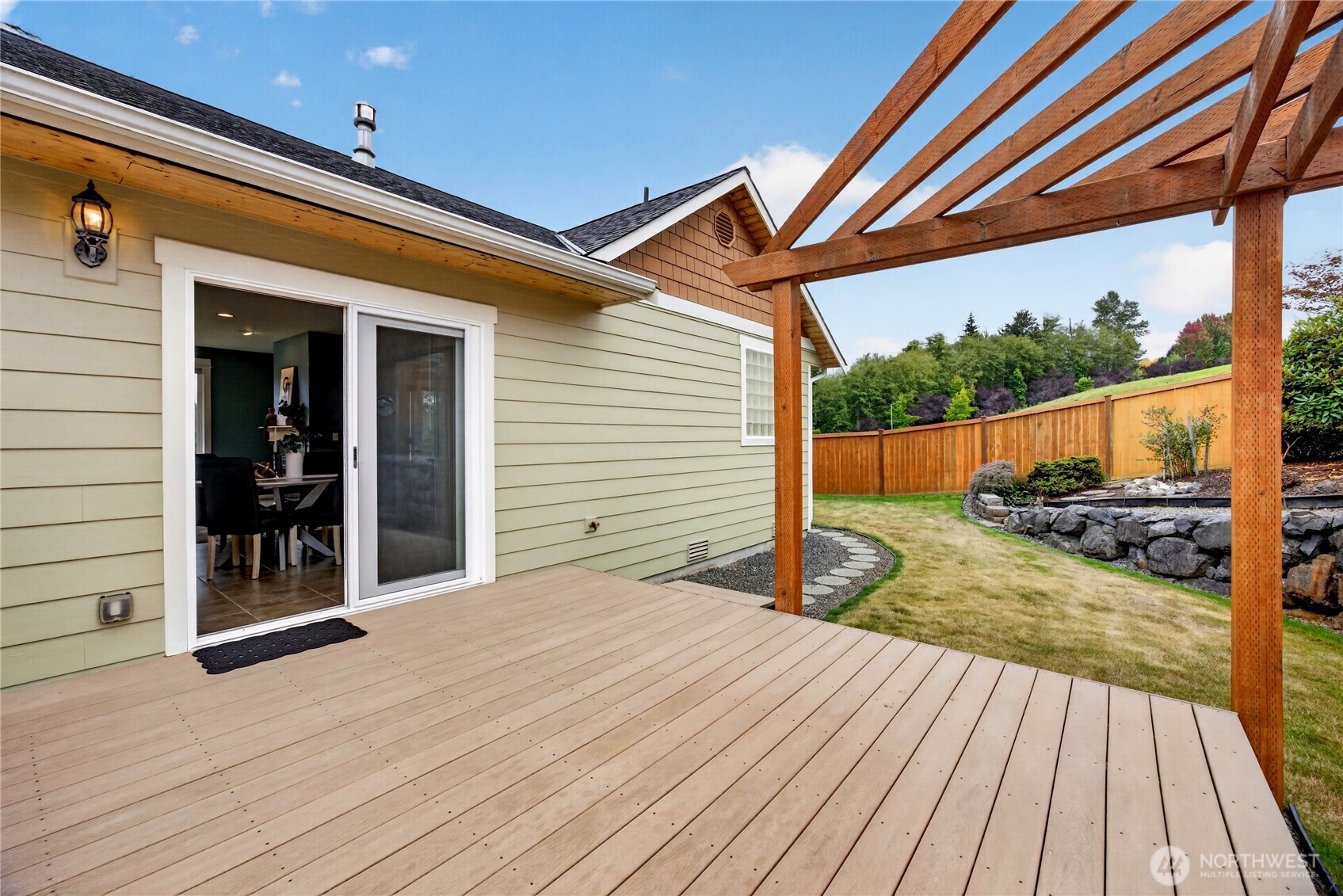 Spacious wood deck beneath a timber pergola beside a fenced lawn and landscaped rock wall.