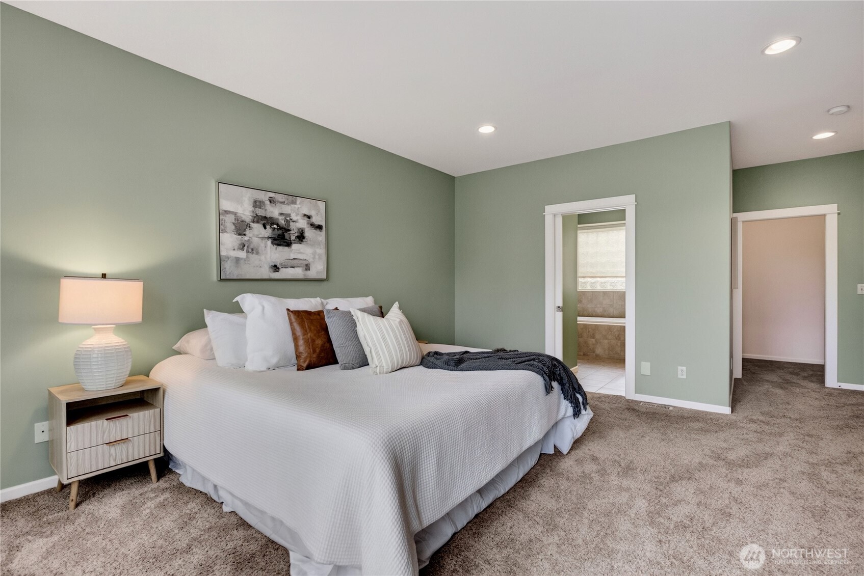 Staged bedroom with a large white bed, sage-green walls, and doorways to a bathroom and adjoining room.