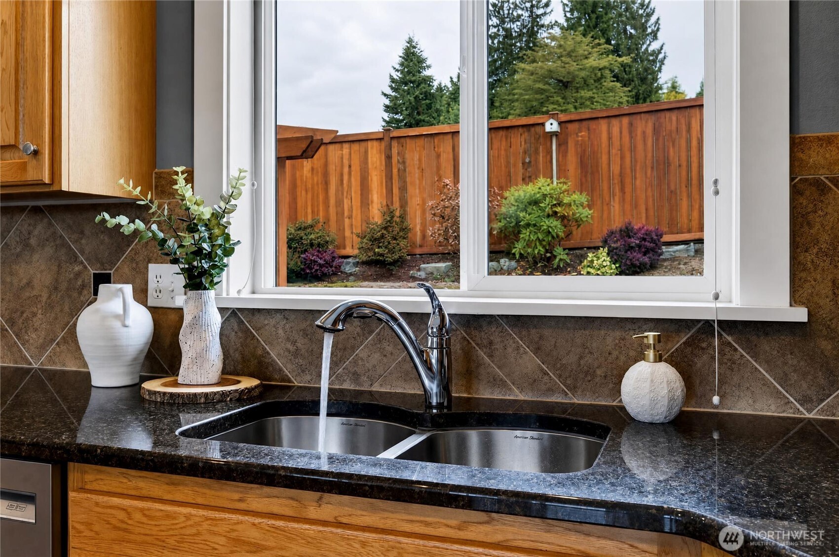 Kitchen sink beneath a window overlooking a landscaped yard, with dark granite counters and wood cabinetry.