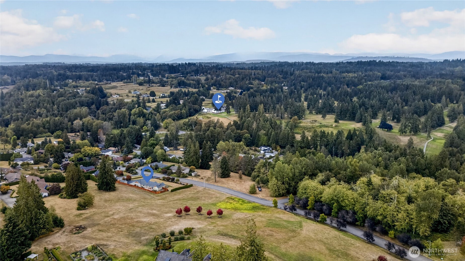 Aerial view of a home beside open fields, with woods and distant mountains beyond.