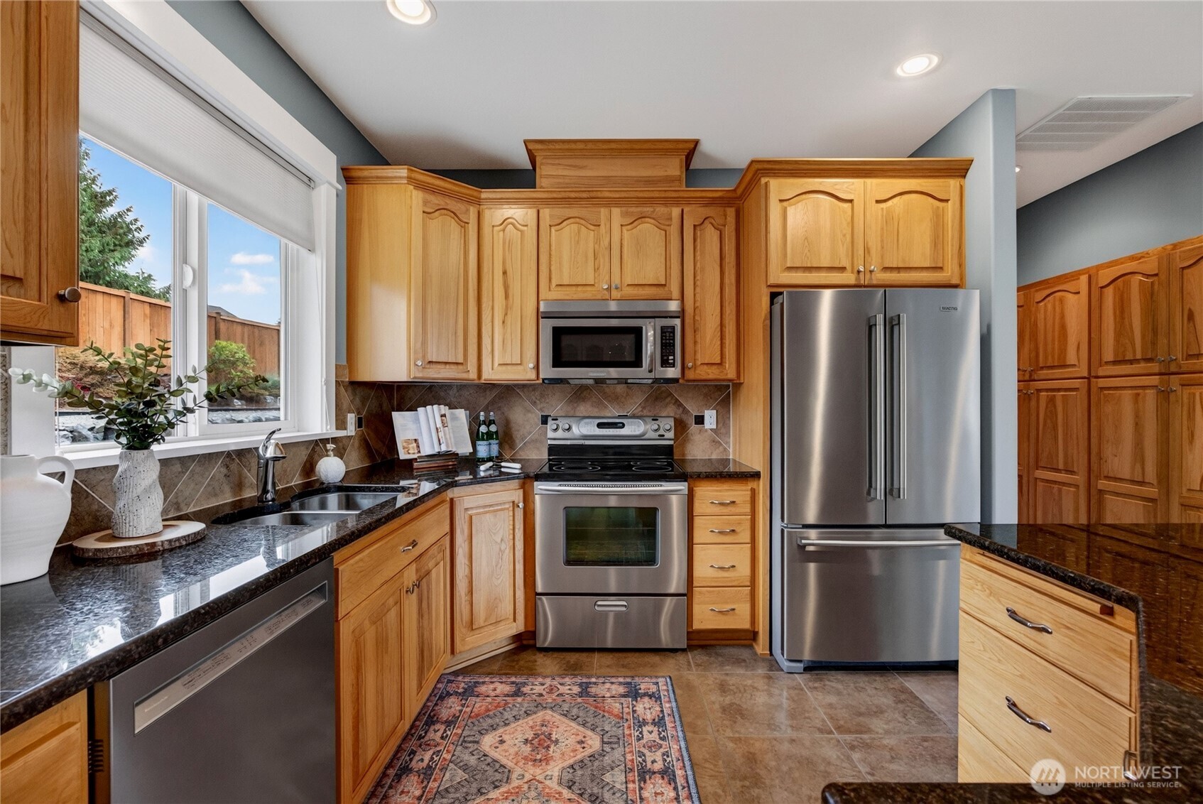Kitchen with honey-colored wood cabinets, dark countertops, stainless steel appliances, and a large window.