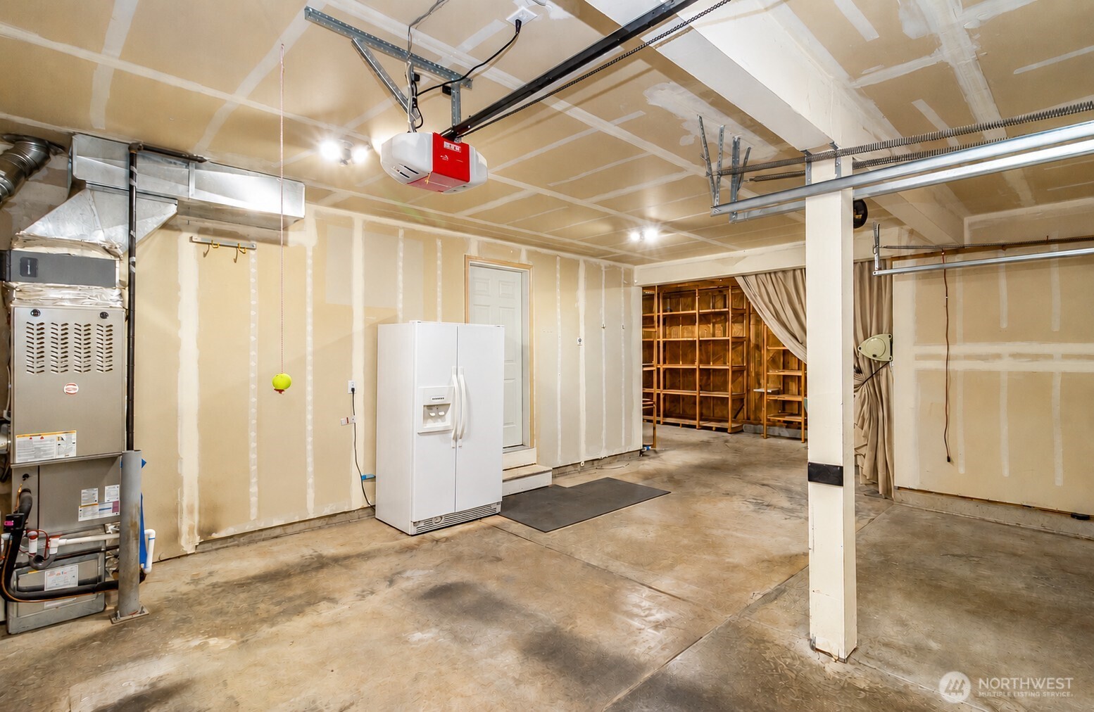 Unfinished garage with exposed drywall, garage door tracks, a furnace, refrigerator, and storage shelves.