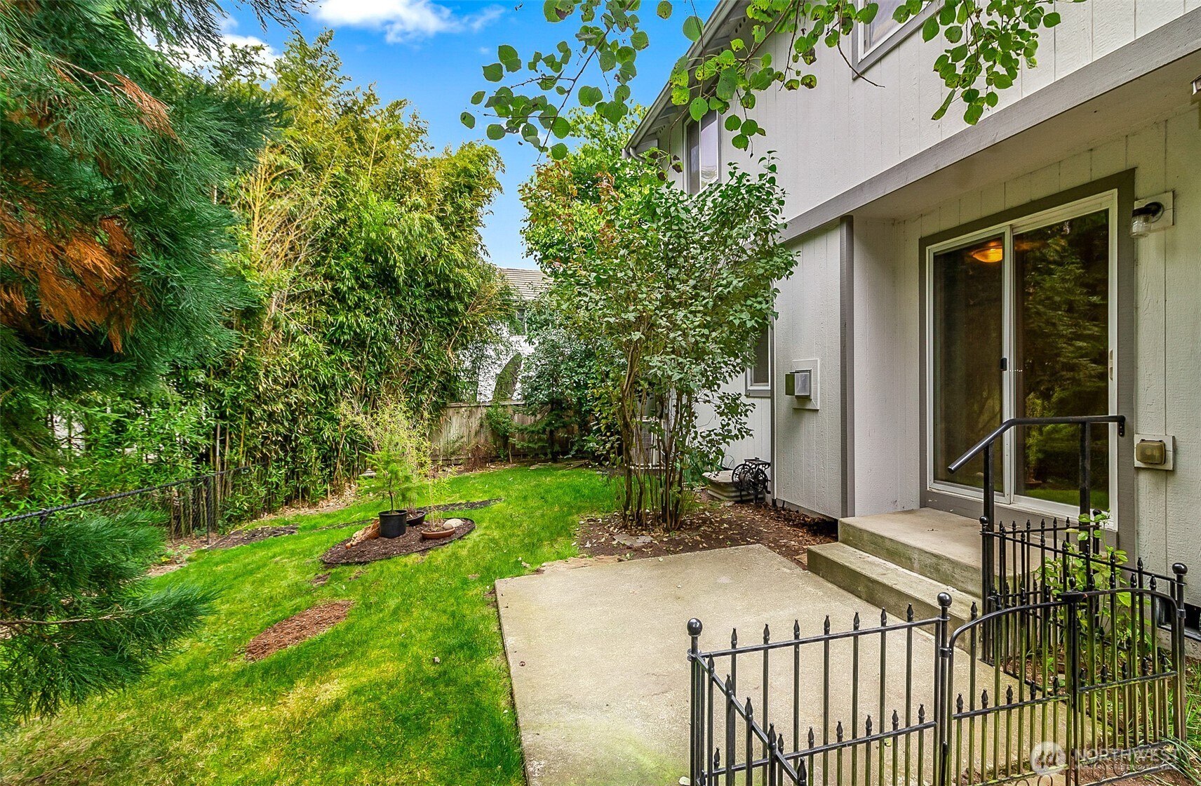 Backyard patio beside the house, with a fenced lawn shaded by mature trees and shrubs.