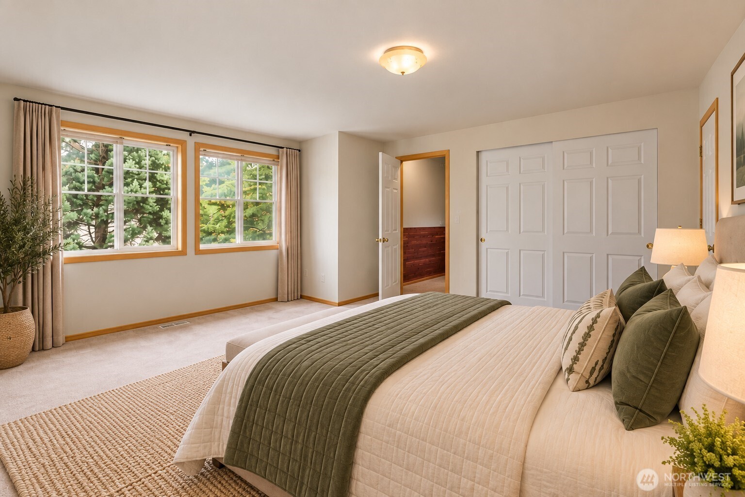 Furnished bedroom with a large bed, olive-green quilt, wood-trimmed windows, and leafy trees outside.