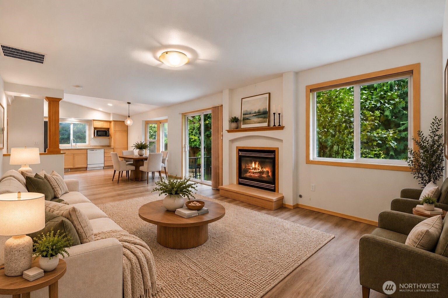 Open-plan living room with a gas fireplace, large leafy windows, and adjoining kitchen and dining area.