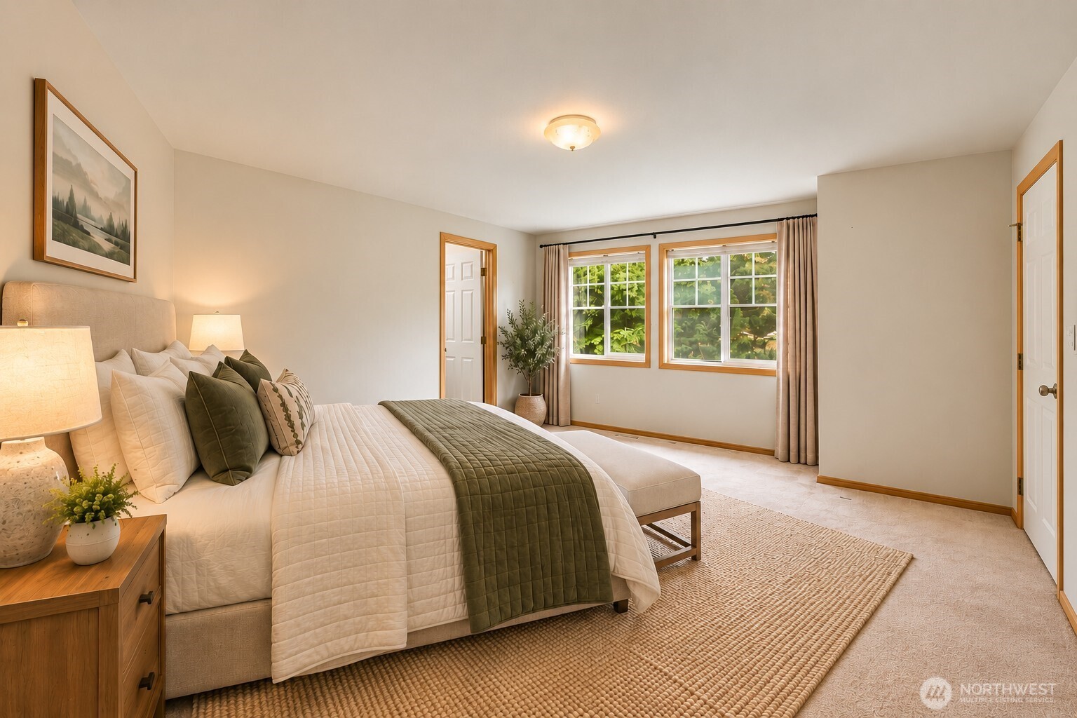 Staged bedroom with a large white bed, olive green quilt, and two curtained windows overlooking greenery.