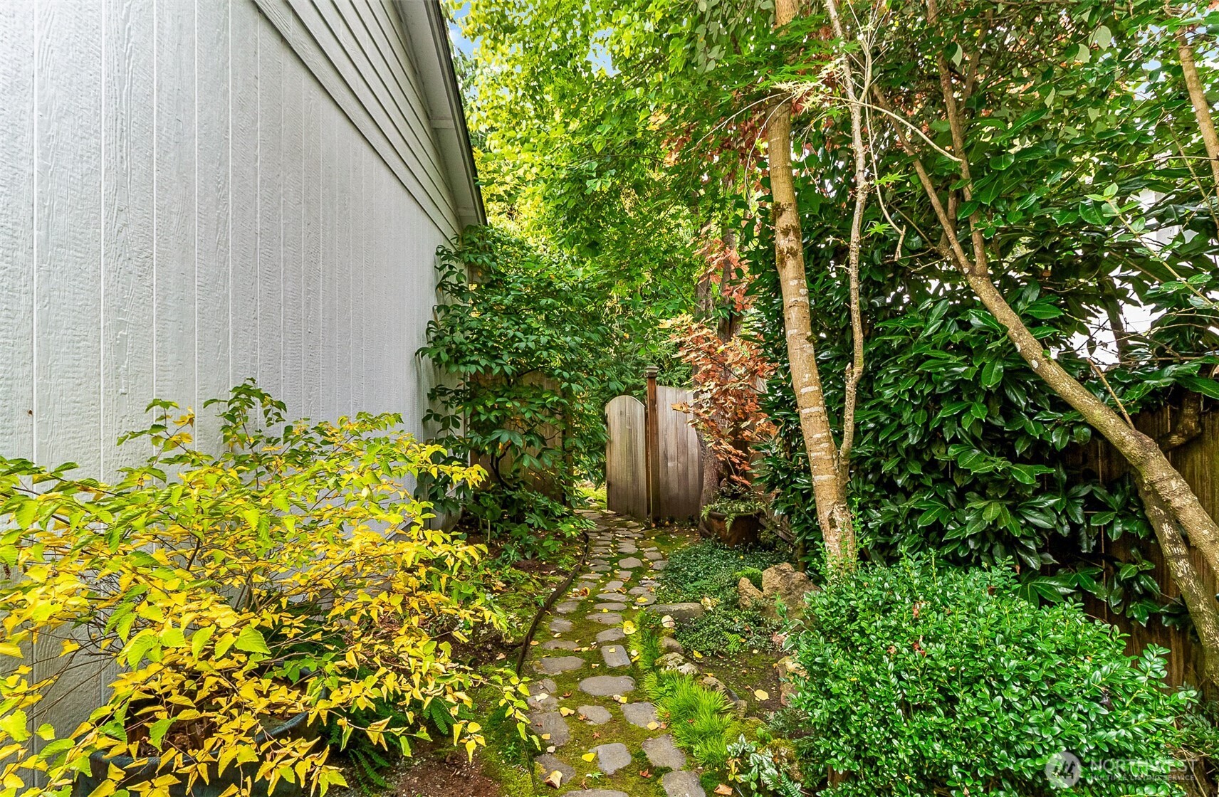 Stone path winds through leafy landscaping beside the house toward a wooden gate.