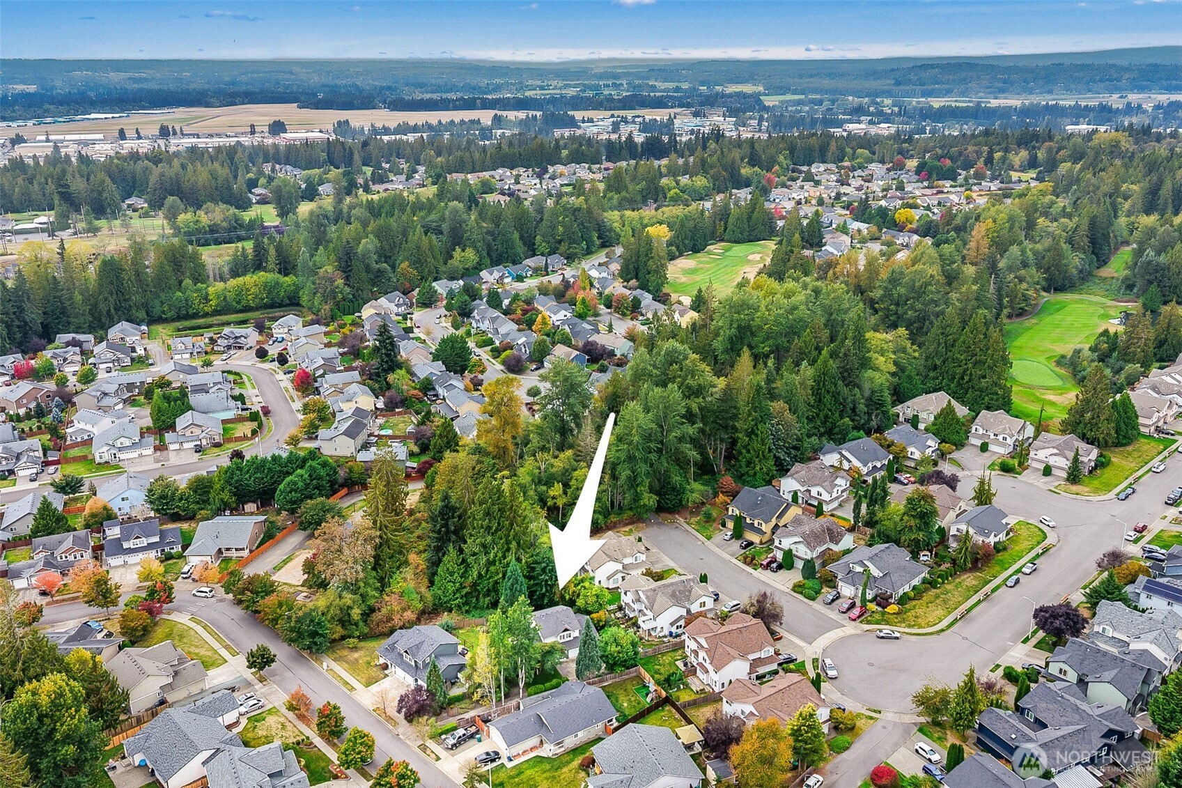 Aerial view of a neighborhood property marked by a white arrow, near wooded areas and a golf course.