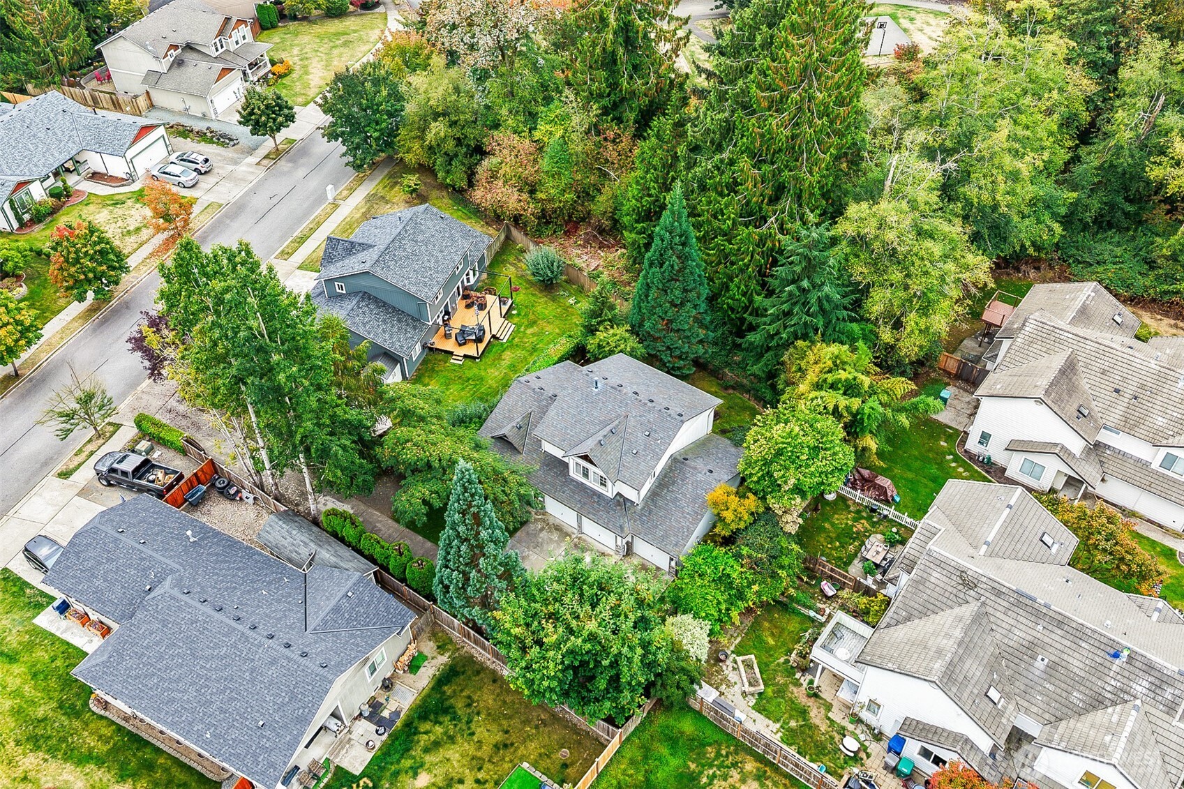 Aerial view of a gray home with a wooden deck amid mature trees and neighboring houses.
