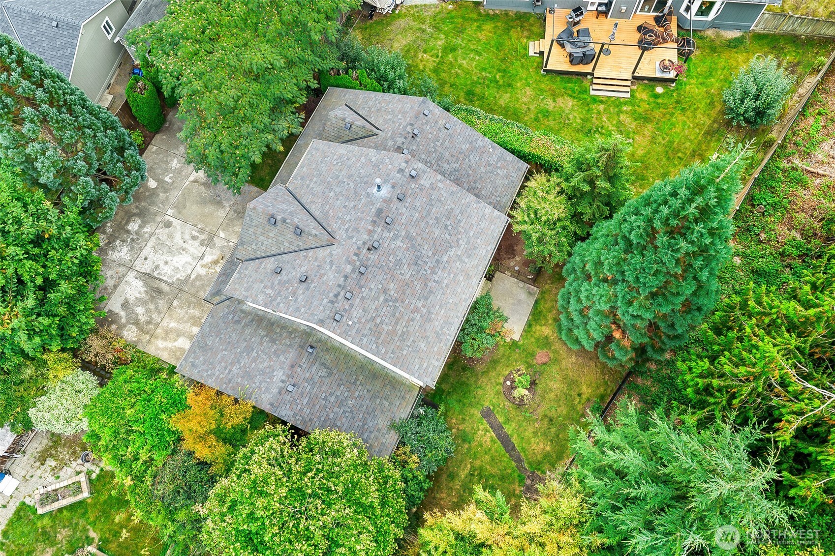Aerial view of a shingle-roofed home surrounded by mature trees, with a lawn, patio, and furnished deck.