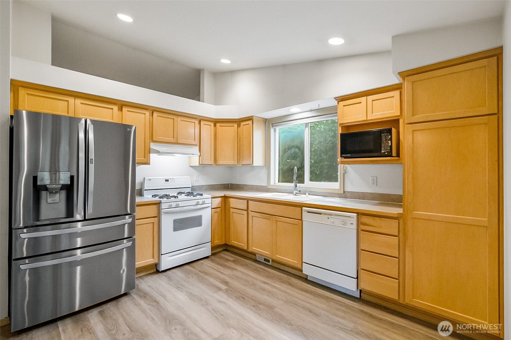 Kitchen with honey-colored cabinets, stainless refrigerator, and a window above the sink.