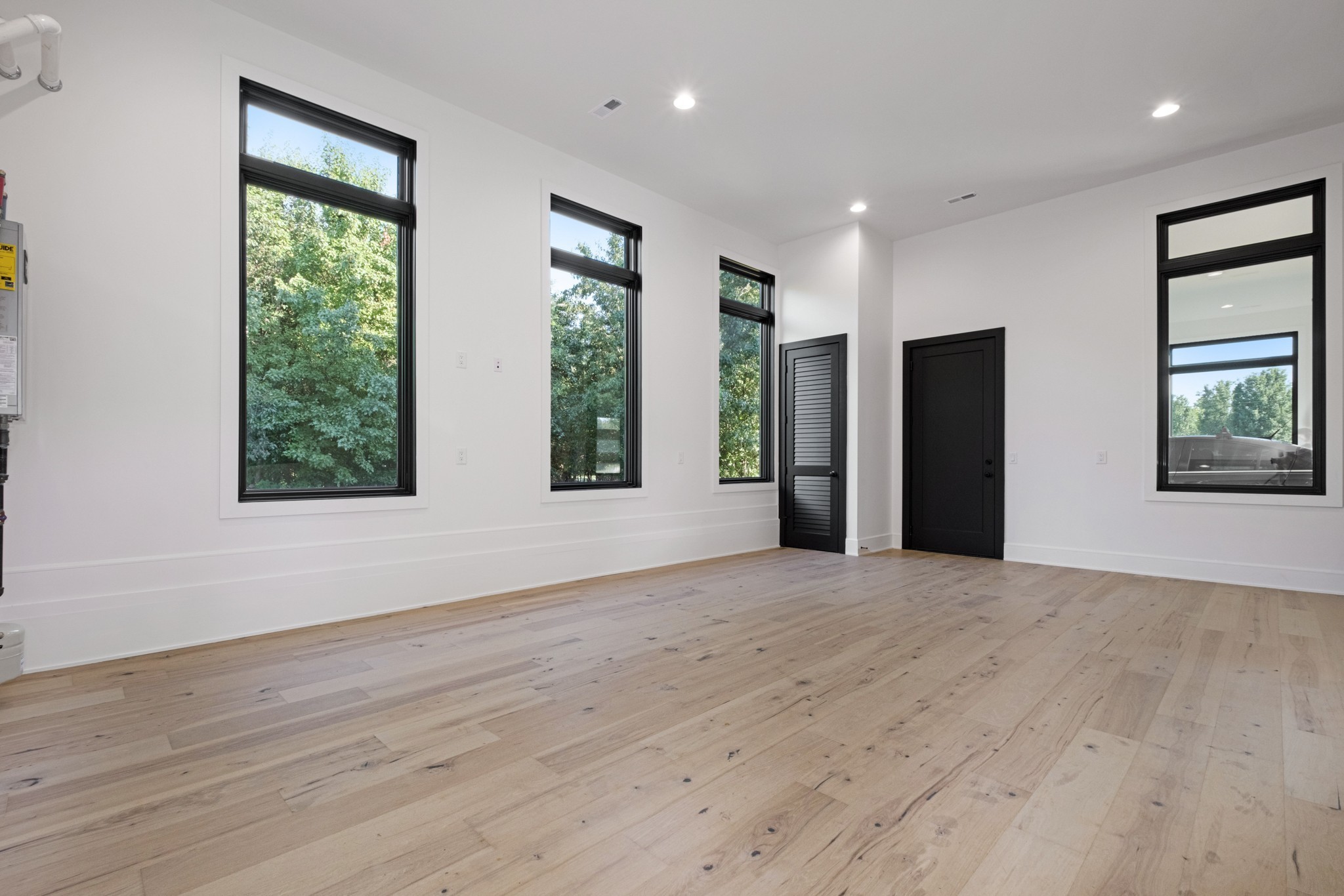 Vacant room with pale hardwood floors, tall black-framed windows, and black doors.