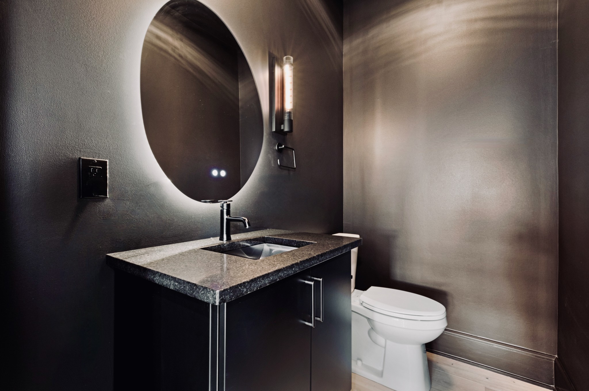 Powder room with a black vanity, speckled stone countertop, and backlit oval mirror.