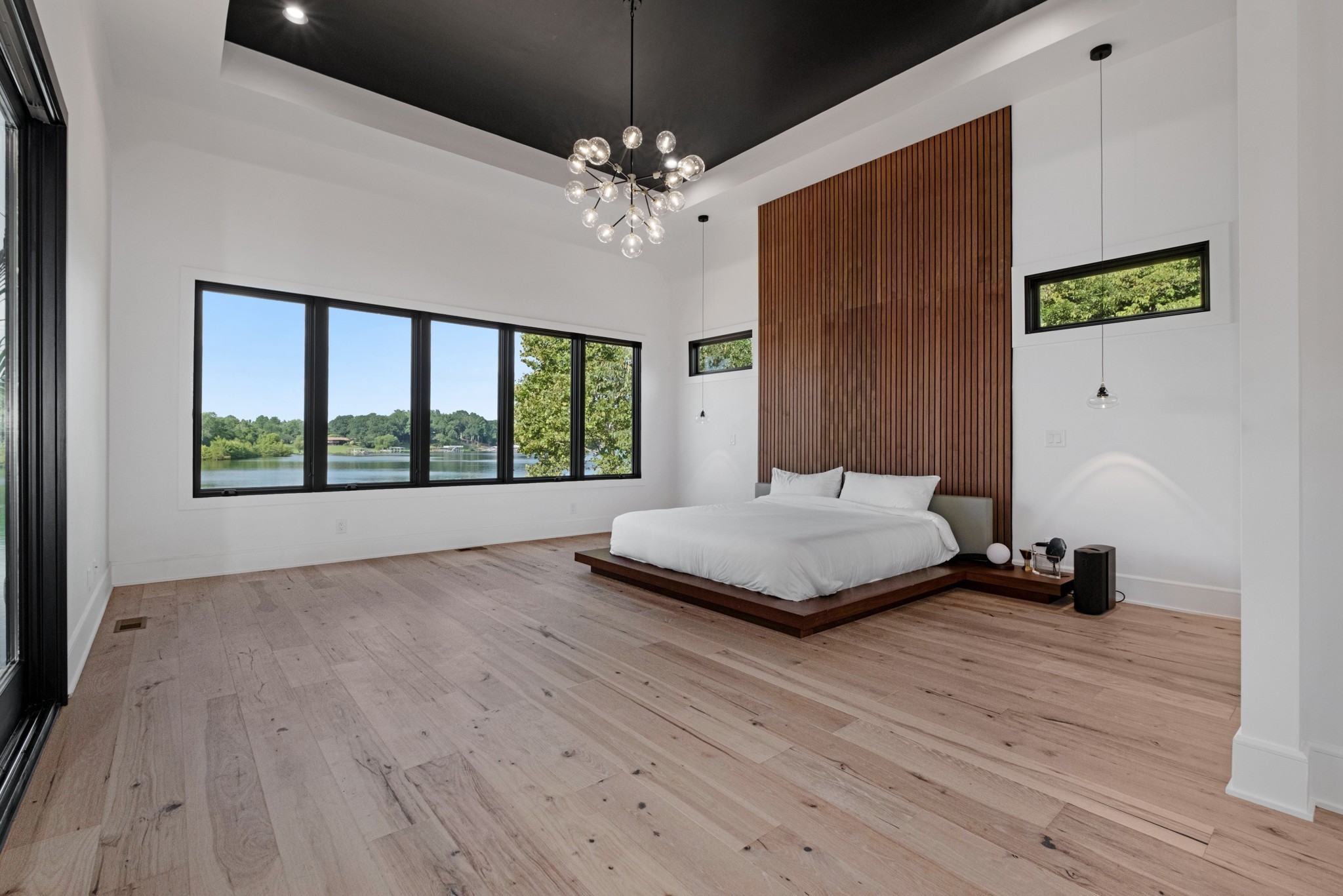 Modern bedroom with a platform bed, wood-slat accent wall, and wide windows overlooking a lake.