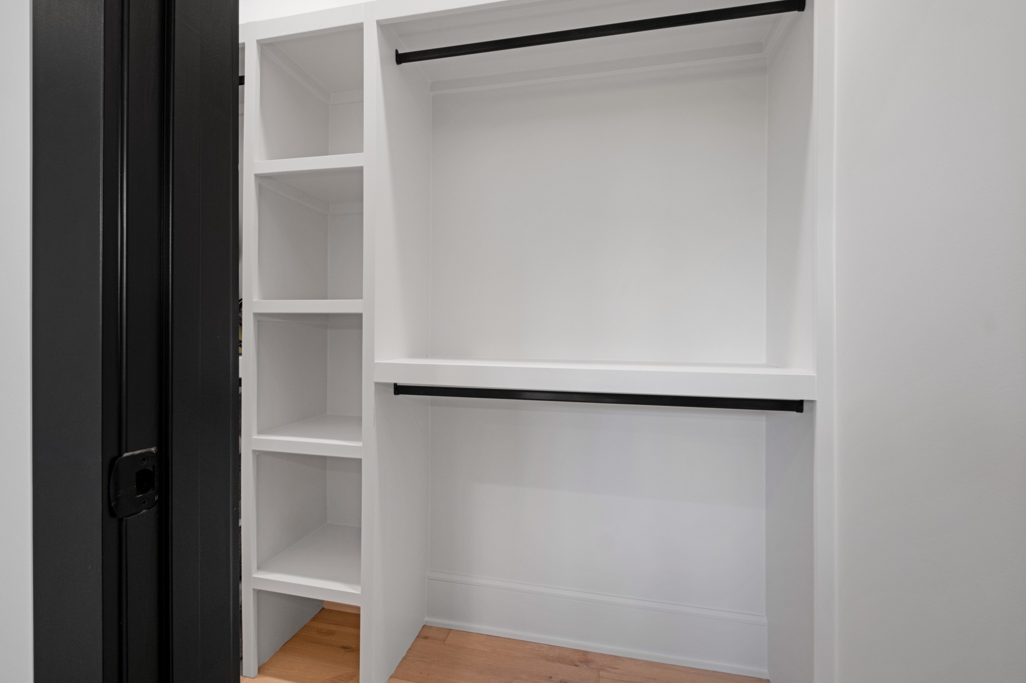 Empty walk-in closet with white built-in shelves, black hanging rods, and a light wood floor.