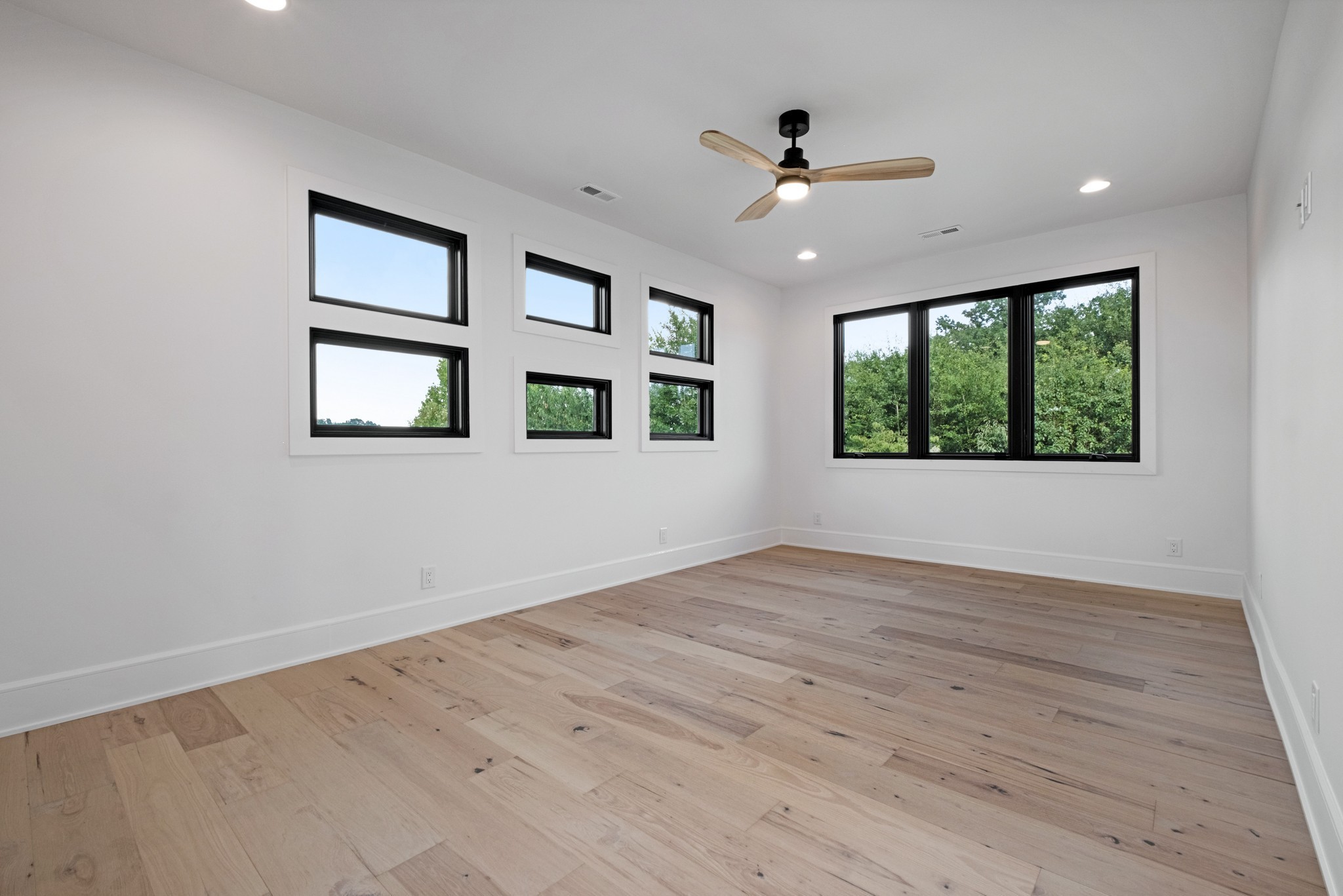 Empty room with pale wood floors, multiple black-framed windows, and a ceiling fan.