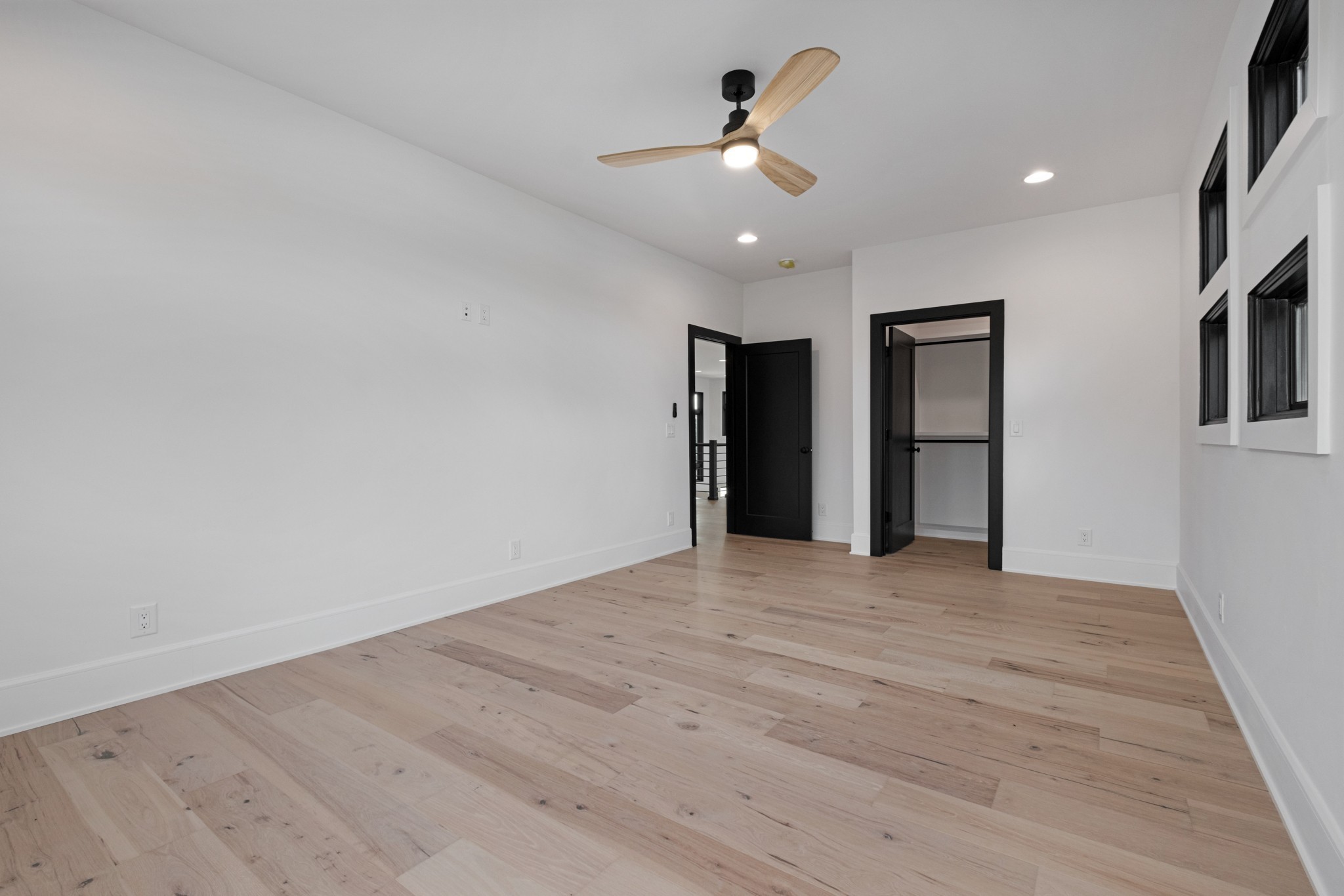Empty room with light wood flooring, black-trimmed doors and windows, and a wood-blade ceiling fan.
