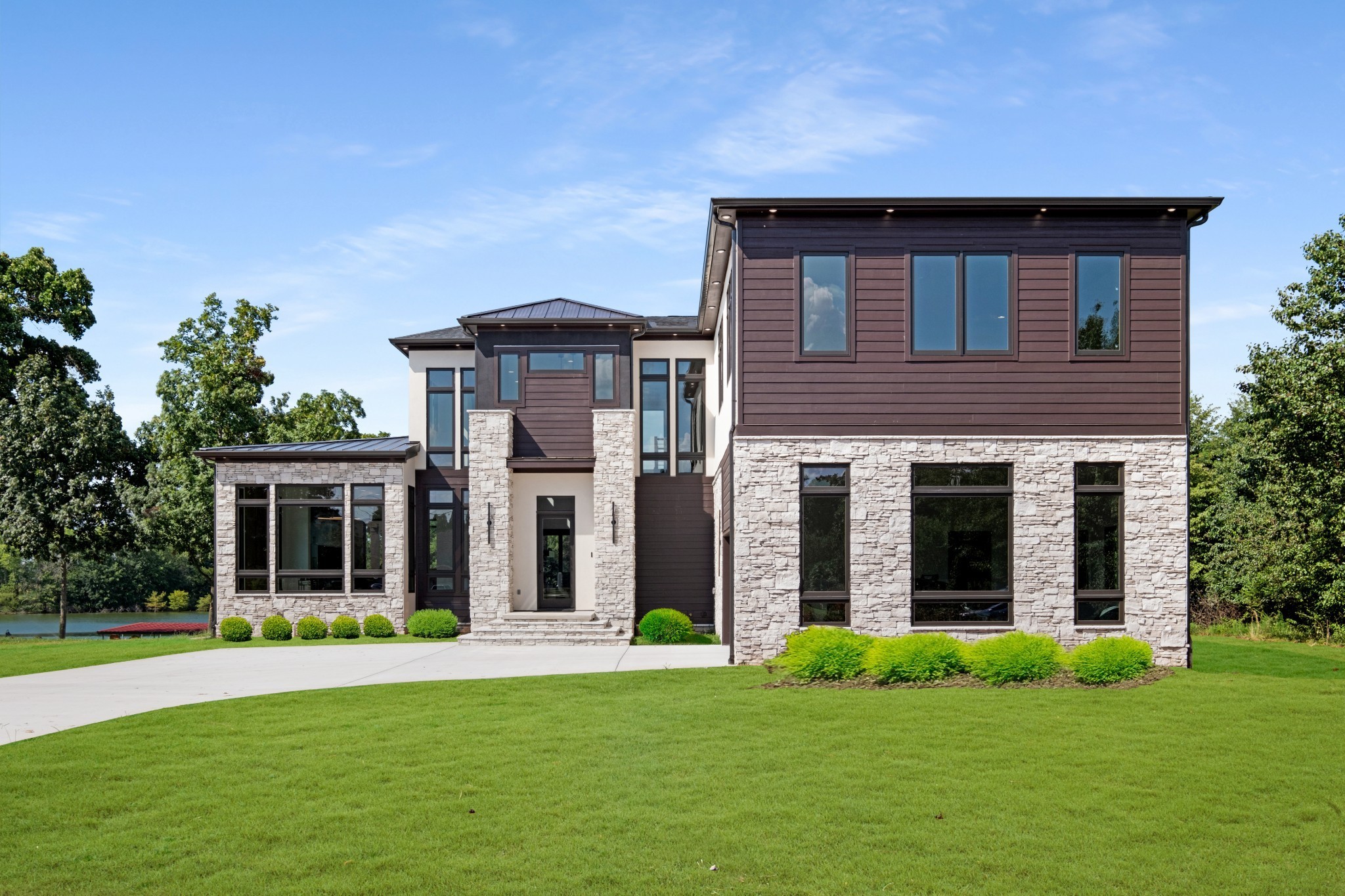 Modern two-story stone-and-siding home with a broad lawn and water visible behind it.