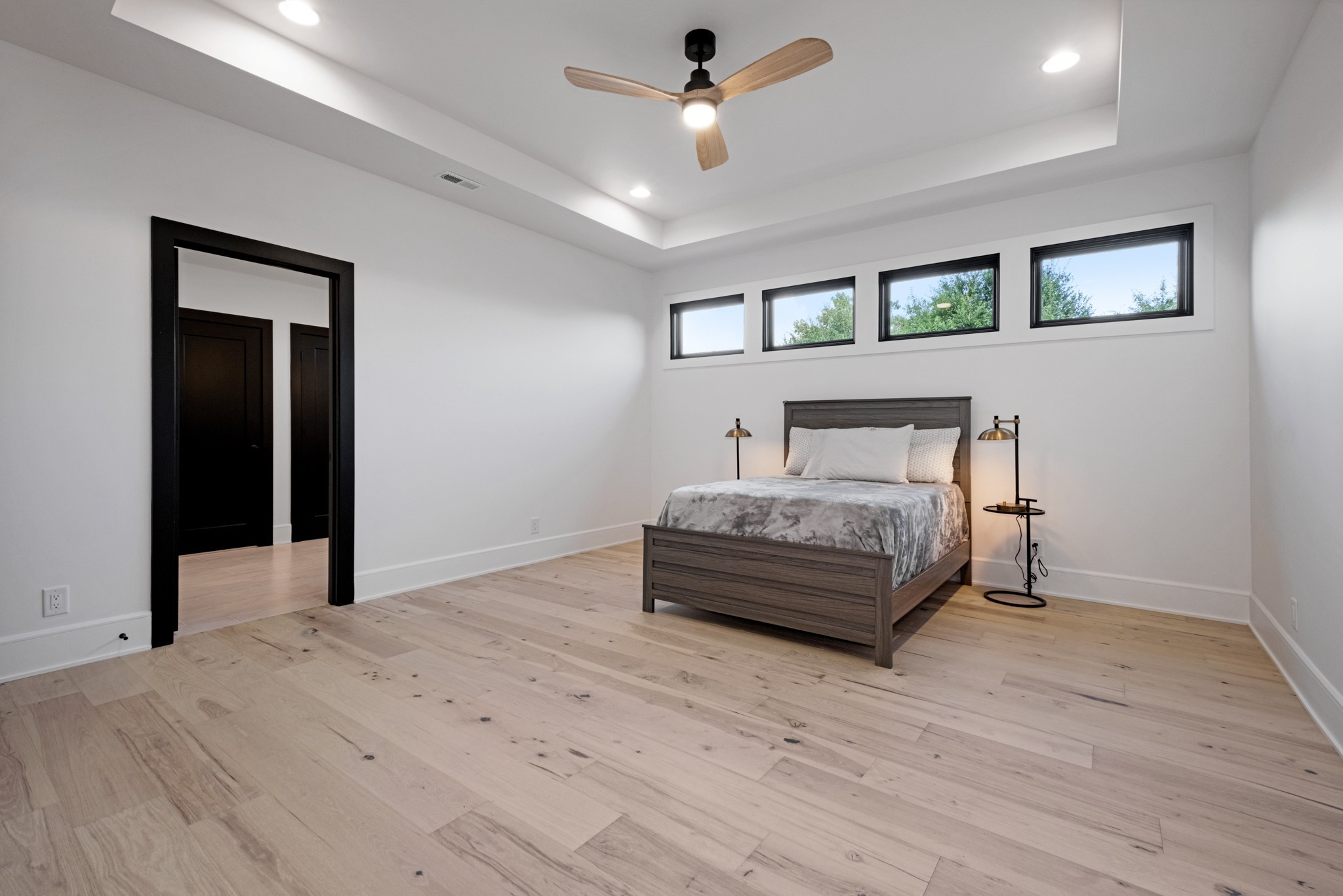 Furnished bedroom with a gray bed, pale wood floors, and four high windows.