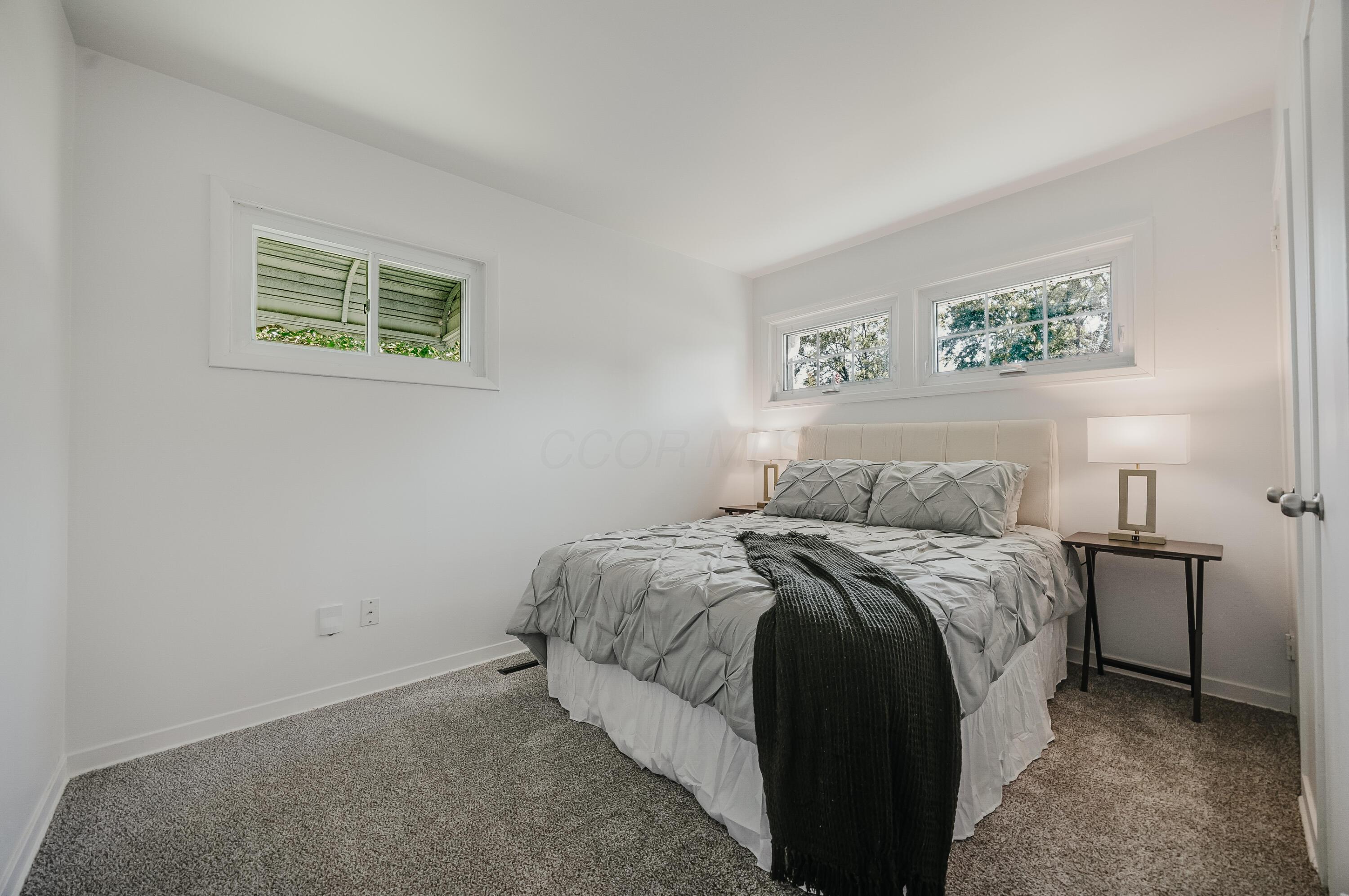Staged bedroom with a gray bed, dark throw, bedside lamps, and high windows.