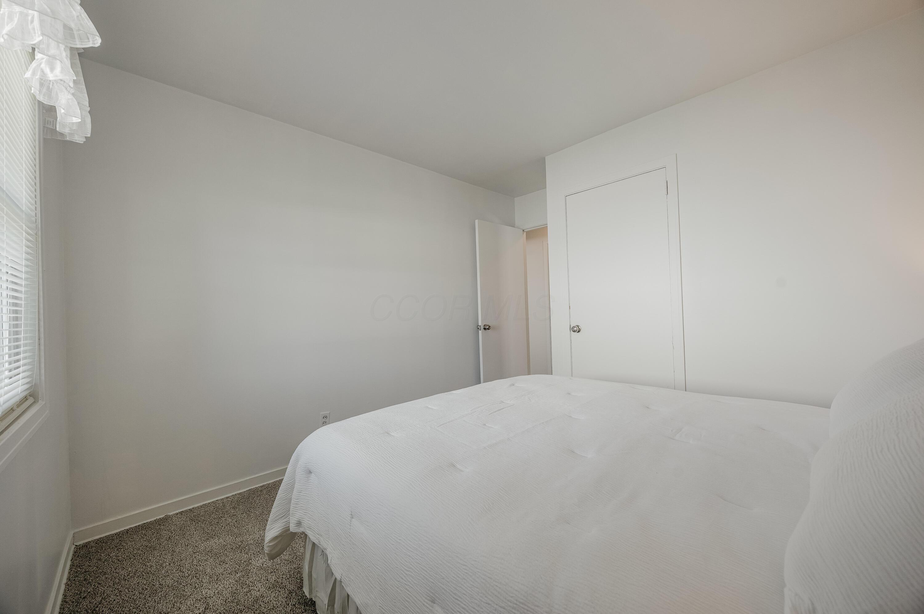 Bedroom with a white bed, beige carpet, and a window with blinds and a ruffled valance.