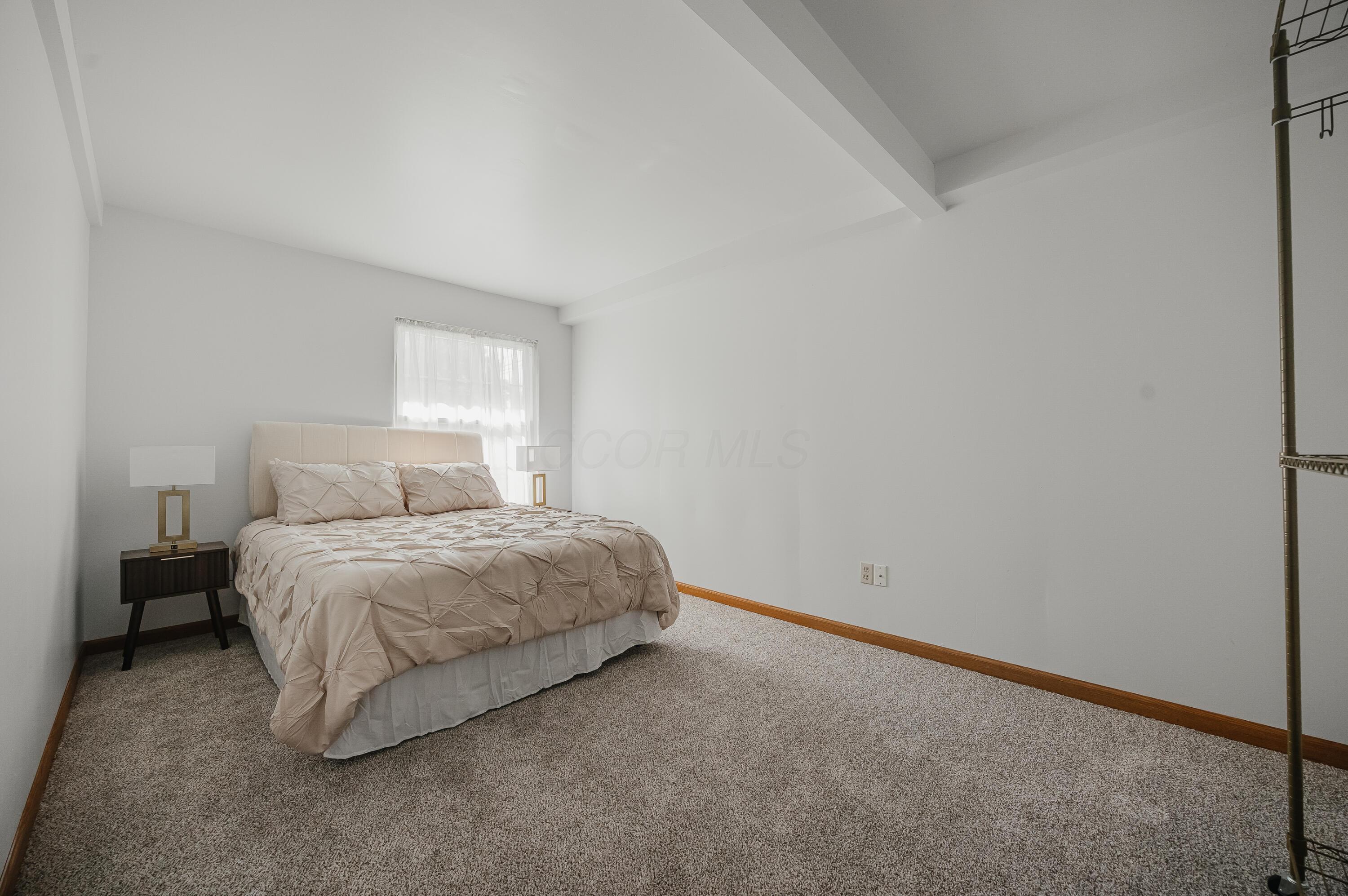 Bedroom with a beige bed, dark bedside table, and broad expanse of gray carpet.